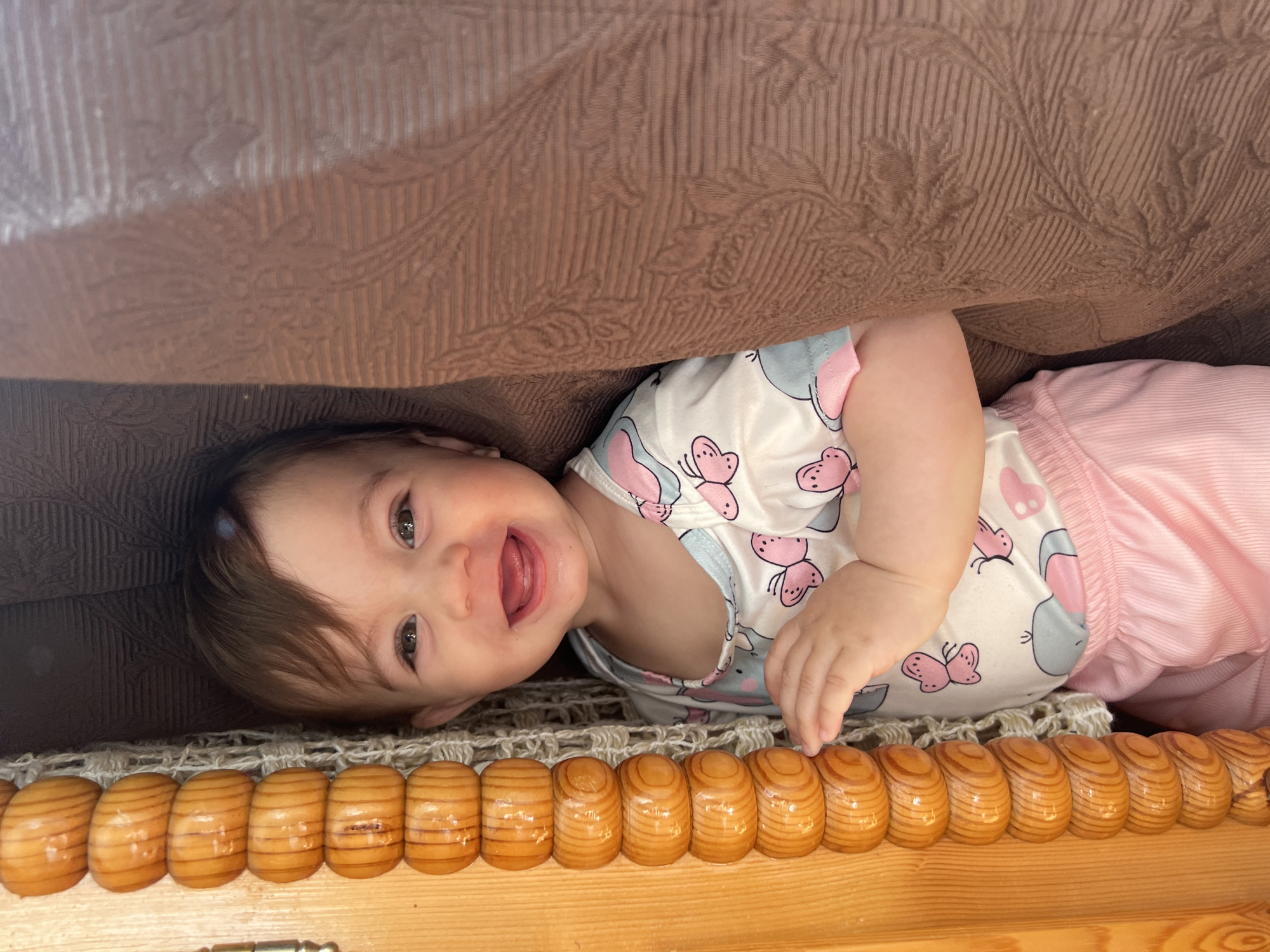 A baby lies on its side, smiling, under a brown textured surface, possibly a couch. The baby is wearing a white shirt with pink butterflies and gray elephants, and pink pants. The surface the baby is lying on features a row of rounded wooden segments with a woven material behind them. The image location is Elena, Bulgaria, however, the surrounding city cannot be seen.