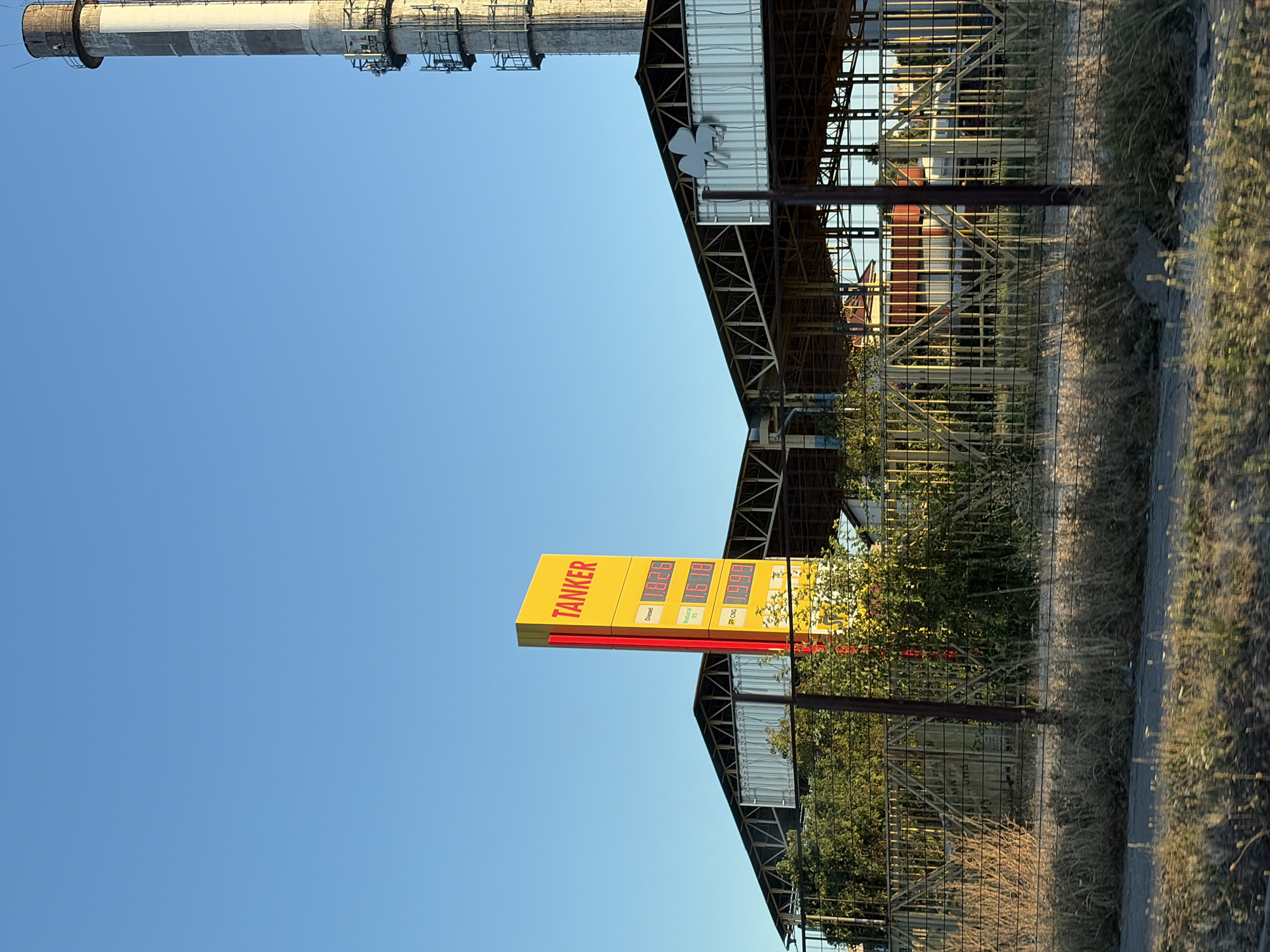 This is a photograph of a gas station in Nitra, Slovakia, on a clear day. A tall, yellow sign with red lettering reading "TANKER" stands prominently in the foreground, displaying fuel prices for Diesel, Natural Gas, and CNG. Behind the sign is a low-slung building with a metal roof supported by a skeletal structure. To the right of the gas station, a tall, industrial smokestack rises against the blue sky, with visible scaffolding and equipment attached to its exterior. A metal fence runs along the perimeter of the gas station, separating it from a grassy area in the foreground. The scene appears to be captured from a moving vehicle, indicated by the motion blur in the foreground grass and the slight blur on the fence.