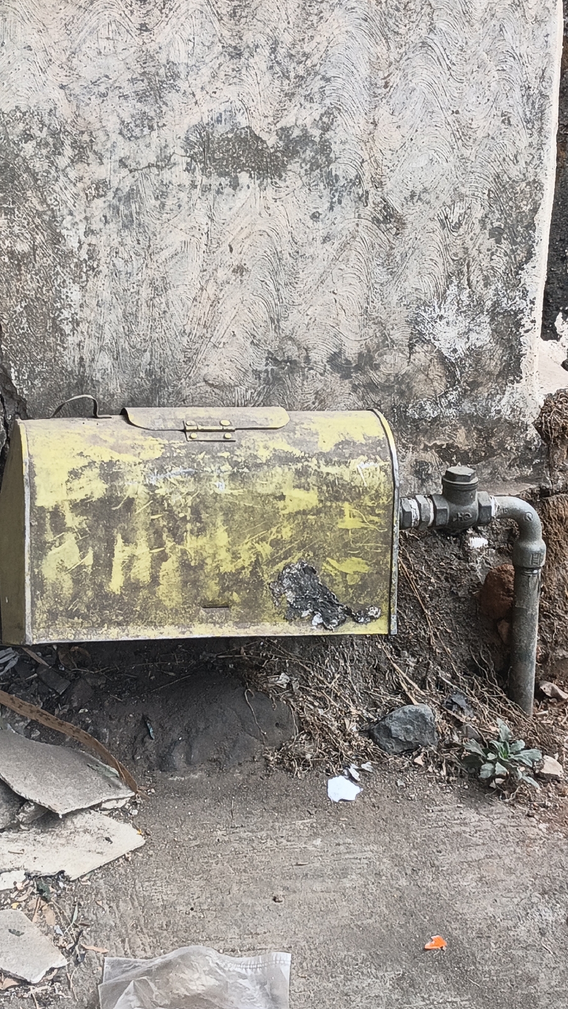 The image depicts an outdoor scene in Dawe, Indonesia, with a focus on a yellow metal utility box mounted on a weathered concrete wall. The box has a hinged lid and appears to be old and somewhat deteriorated, with visible rust and peeling paint, particularly on its right side. A pipe with a valve emerges from the right side of the box and connects to a larger vertical pipe. The wall behind the box is textured and stained, showing a wavy pattern from what appears to be a previous plaster application or damage. The ground in front of the wall is a mixture of dirt, dry vegetation, and debris, including broken concrete fragments, small stones, and pieces of white paper. A small green plant is visible growing in the dirt near the right side of the scene. The overall impression is one of an aged and somewhat neglected urban utility area.