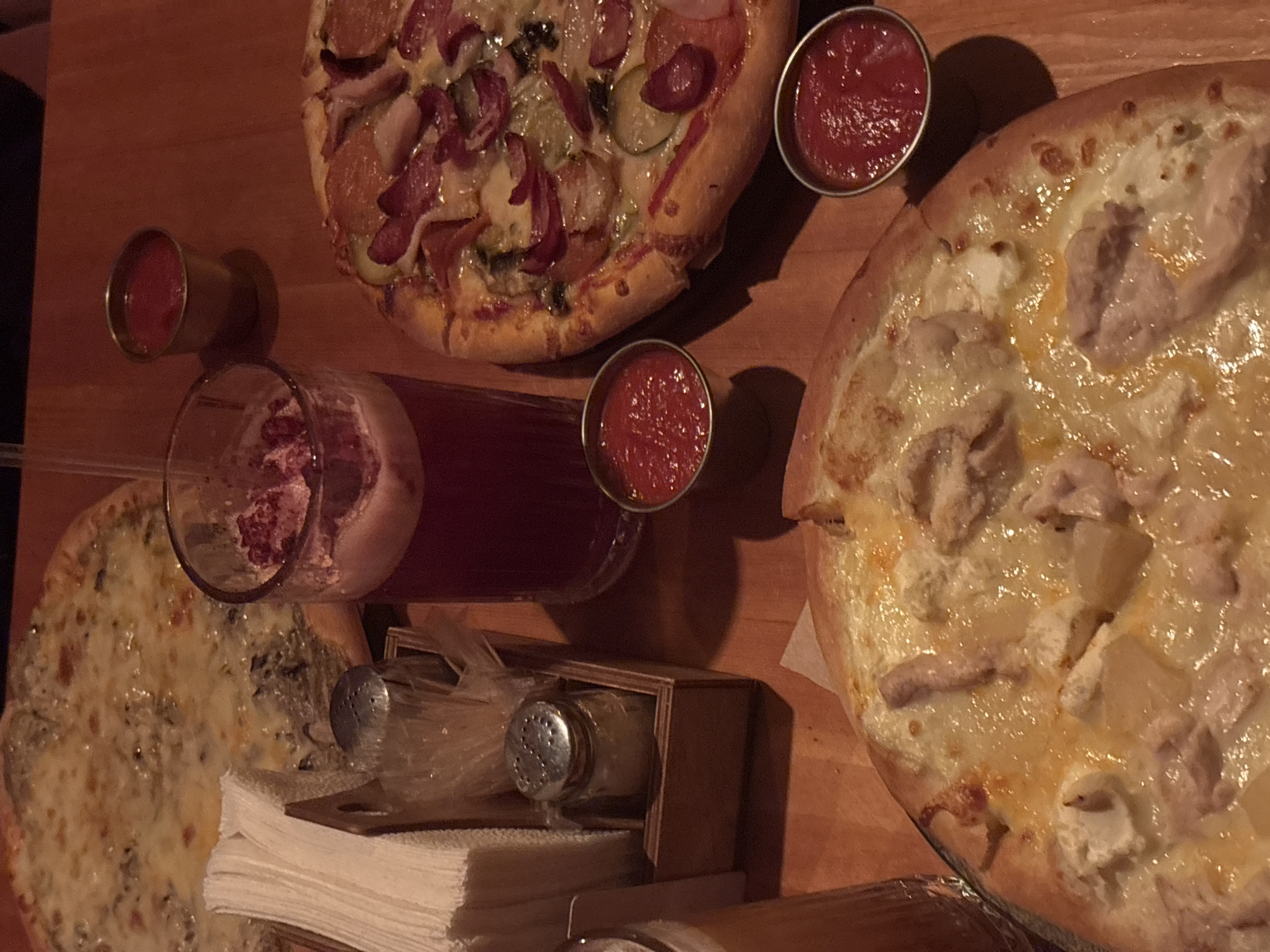 The image displays an overhead view of a dining table laden with food and drinks, suggesting a casual meal during the evening or night, indicated by the warm, dim lighting and visible shadows. The setting is likely a restaurant or bar in Perm, Russia.
The primary subjects are three pizzas, each with distinct toppings. The pizza at the top features a red sauce base with slices of pepperoni or salami, red onion, and what appear to be pickles. To its right, another pizza has a white sauce base, topped with melted cheese, chunks of chicken, and pineapple. Partially visible on the bottom left is a third pizza, also with a white sauce base, covered in cheese and mushrooms, possibly a four-cheese or mushroom pizza.
Accompanying the food are several items. Three small metallic cups hold a bright red sauce, likely tomato sauce or ketchup. Two clear glasses contain a dark red liquid, possibly a berry drink or dark beverage, with one glass showing reddish-purple sediment at the bottom. A stack of white paper napkins rests on the wooden table, beside a small wooden caddy that holds salt and pepper shakers, each individually wrapped in plastic. The table itself is made of warm-toned wood, contributing to a cozy and inviting atmosphere. No people are visible in the frame, and there is no visible text.