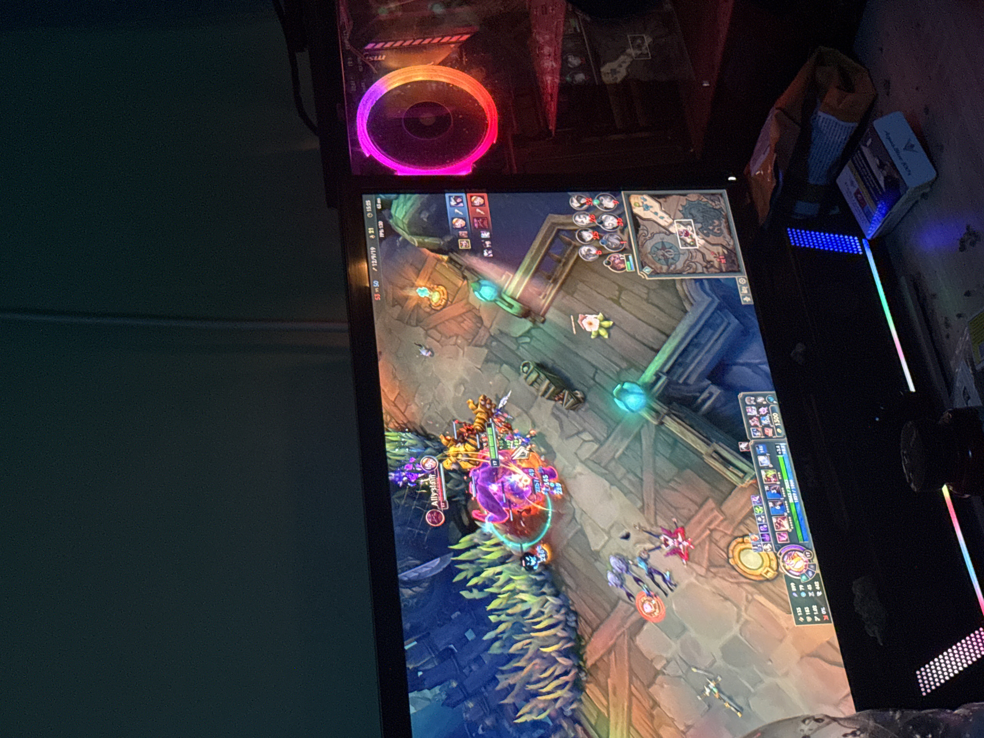 Here is a detailed description of the image:
The image shows a desktop computer playing the game League of Legends. The game is in the middle of a team fight, with champions from both teams battling near a nexus. In the upper left corner, a pink/red illuminated fan from the tower is visible. To the right of the screen are various peripherals, including what looks to be a snack bag and several boxes. The game display shows details like player stats, time, and FPS.