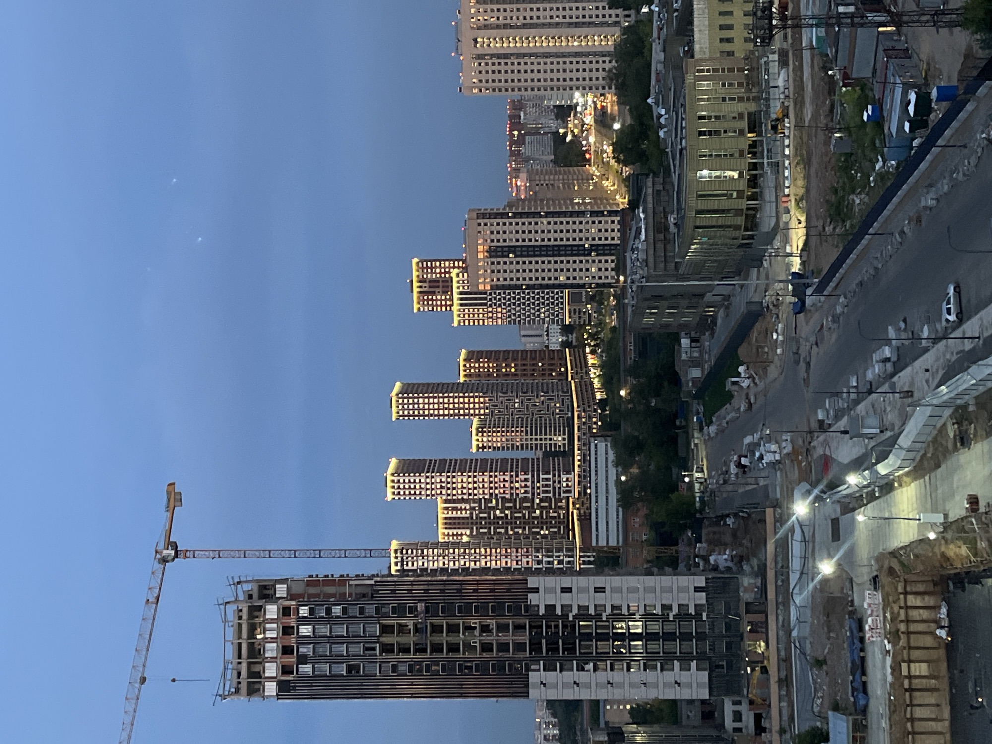 The image captures a twilight view of a city under construction, specifically in the area of Tarasovka, Russia. A large construction crane dominates the left side of the frame, positioned next to a partially built skyscraper. Several other modern high-rise buildings, some illuminated and appearing to be nearing completion, are visible in the background, suggesting a new development area. The foreground shows ongoing construction work, with bare ground, trenches, and stacks of building materials evident. A road winds through the scene, with streetlights illuminating the path. The sky is a deep blue, indicative of dusk, and a few faint lights are visible in the distance, possibly from aircraft or celestial bodies. No people are clearly visible in the image, and the scene is primarily focused on the architectural and infrastructural elements of the developing urban landscape.