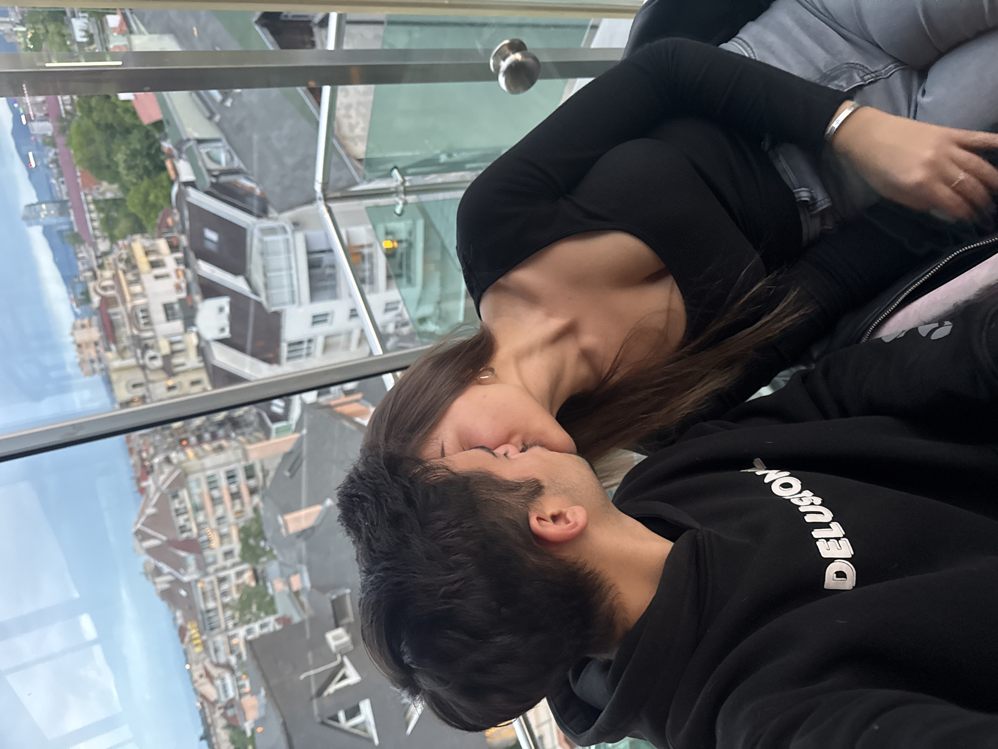 A photograph shows a woman and a man kissing. They are indoors, next to a glass window. Outside the window is a cityscape. The woman is wearing a black long-sleeved shirt and gray jeans. The man is wearing a black hooded sweatshirt with the word "DELUSION" printed vertically on the front. The city outside the window appears to be Sofia, Bulgaria, judging by the architectural style of the buildings.