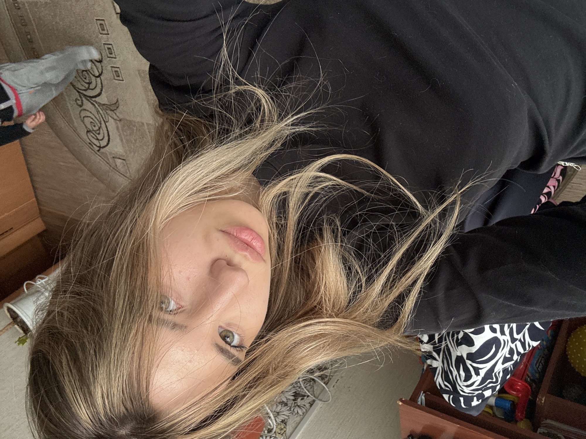 A close-up, selfie-style shot shows a young woman with blonde hair and light green eyes. She is wearing a black long-sleeved shirt. Her face is turned slightly to the right, with her mouth slightly open, revealing pink lips. The background is blurred but includes what appears to be a rug with a swirling pattern and a person wearing grey sweatpants and a dark jacket with red accents walking on it. To the left, a shelf with various items, including a black and white patterned fabric and some toys, is visible. The overall environment suggests an indoor setting, likely a residential room in Noviy, Russia.