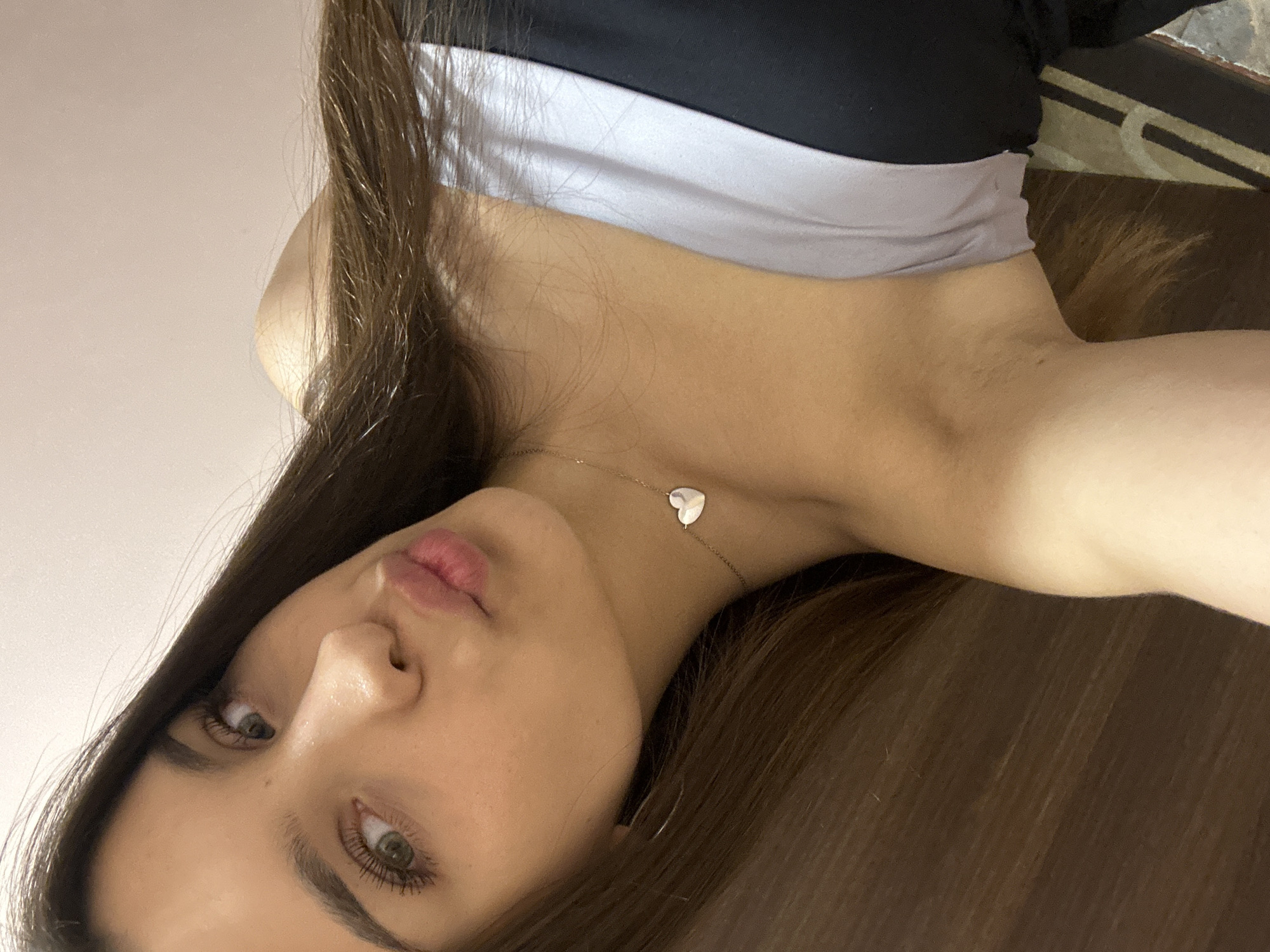 A young woman with long, dark brown hair and green eyes, wearing a strapless top, is posing for a selfie. She is leaning her head to her left, her lips pursed in a kiss. A small, silver heart-shaped pendant is visible on a delicate chain around her neck. The background consists of a plain, light-colored wall. The image does not contain information about the city of Shumen, Bulgaria, or any specific environmental details that would allow for its localization to that city.