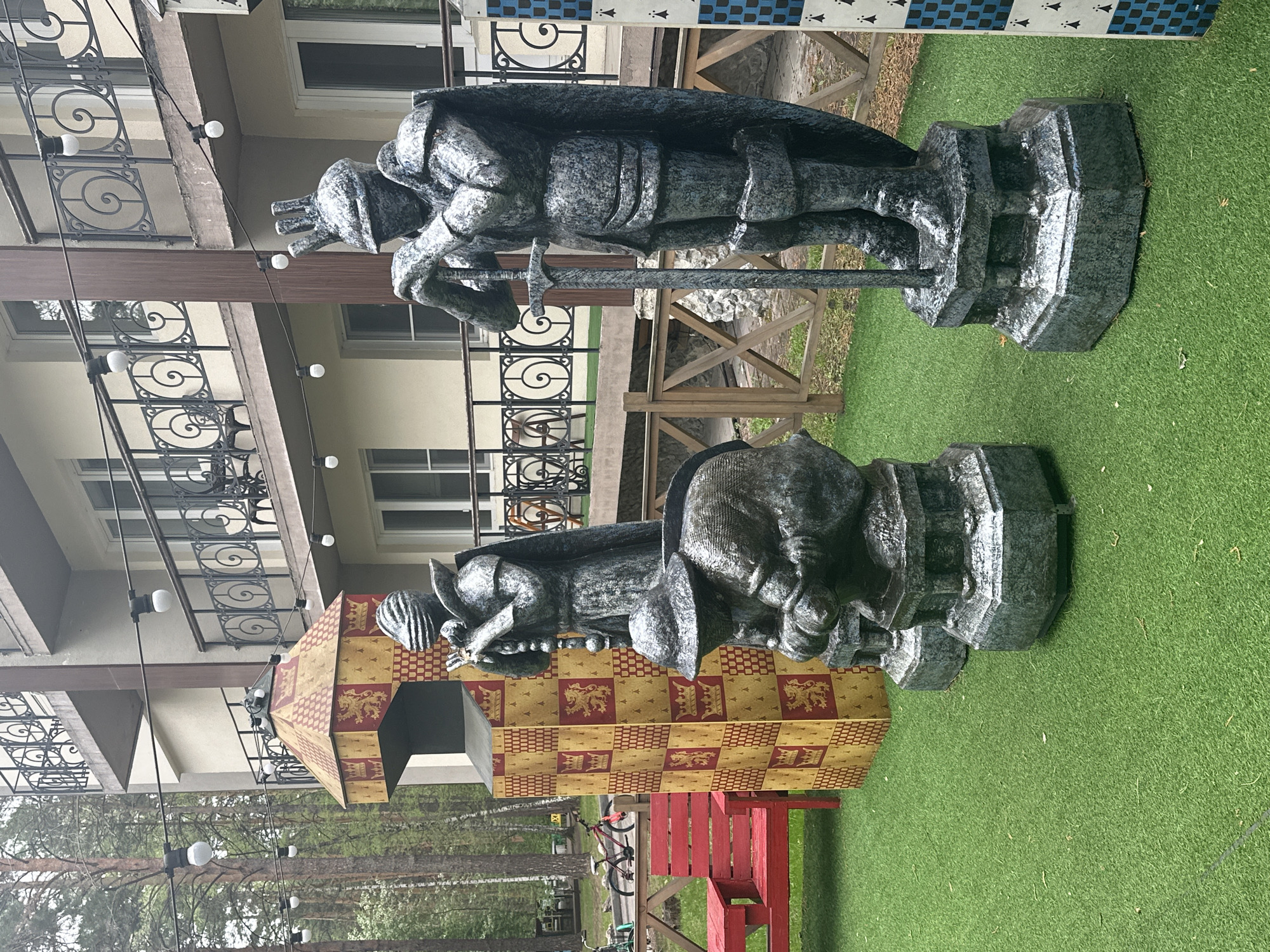 The image shows two large statues of medieval knights made of a gray, stone-like material. One statue is standing, holding a sword and looking down, while the other is bent over. Behind the statues is a building with a cream-colored exterior and decorative wrought-iron balconies. To the left of the statues is a structure decorated with a gold and red checked pattern, possibly representing Gryffindor House colors from the Harry Potter series. A red picnic table is also visible. The statues are on green artificial grass. The location is likely in the city of Berdsk municipality, Russia. The environment seems to be an outdoor recreational area or theme park, based on the presence of the statues and the decorated structure. There are trees in the background, indicating an outdoor setting.