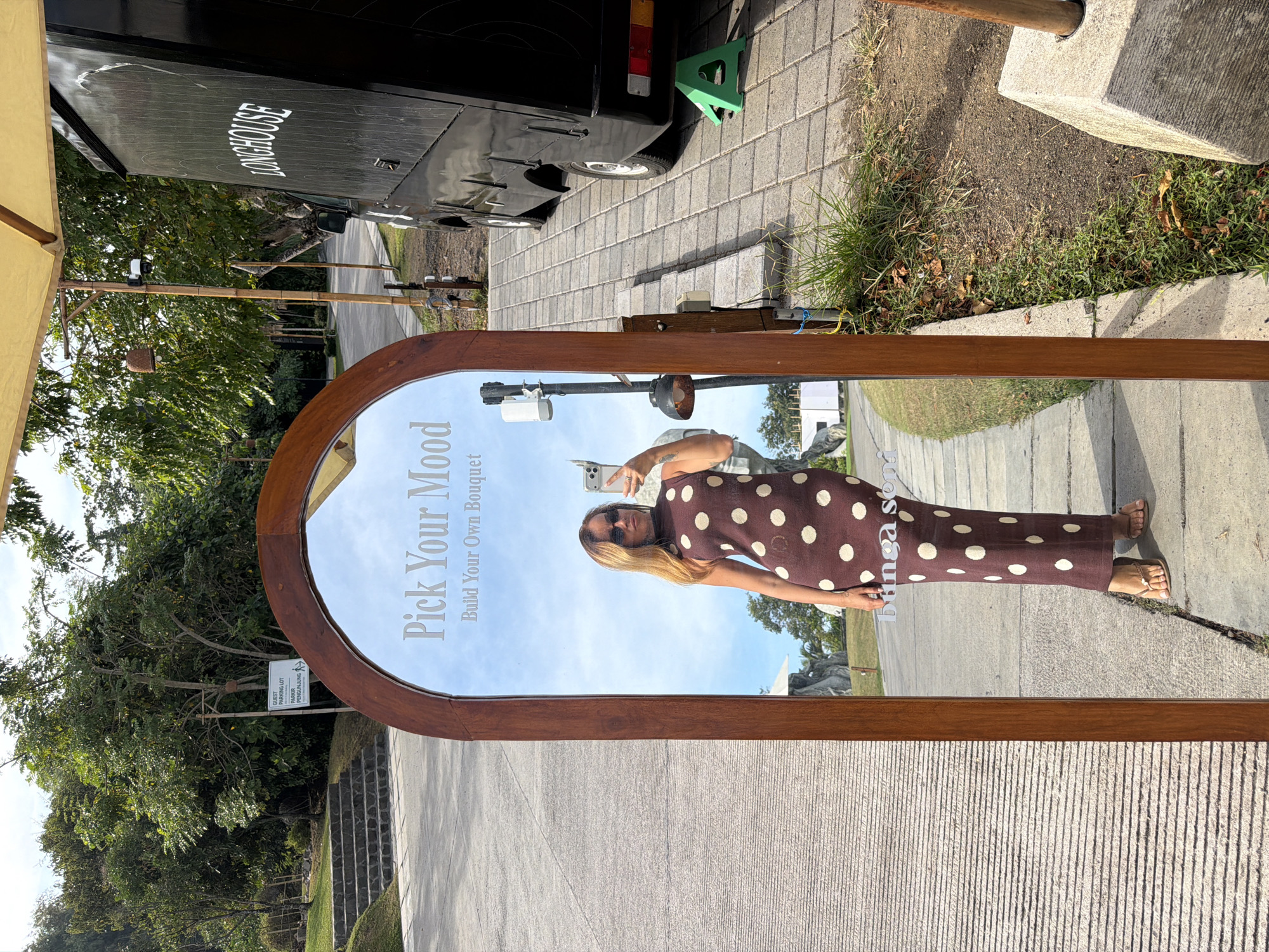 A woman in a polka-dot dress takes a mirror selfie outdoors. She stands on a paved pathway in front of a large arched mirror that reflects her and the sky. The mirror has text that reads "Pick Your Mood" and "Build Your Own Bouquet." To her left, there are grassy slopes and stairs. To her right, a black food truck labeled "LONGHOUSE" is parked. The environment suggests a sunny day with trees and foliage visible in the background. The scene is consistent with an outdoor cafe or marketplace in a city like Kediri, Indonesia, known for its agricultural and natural beauty.