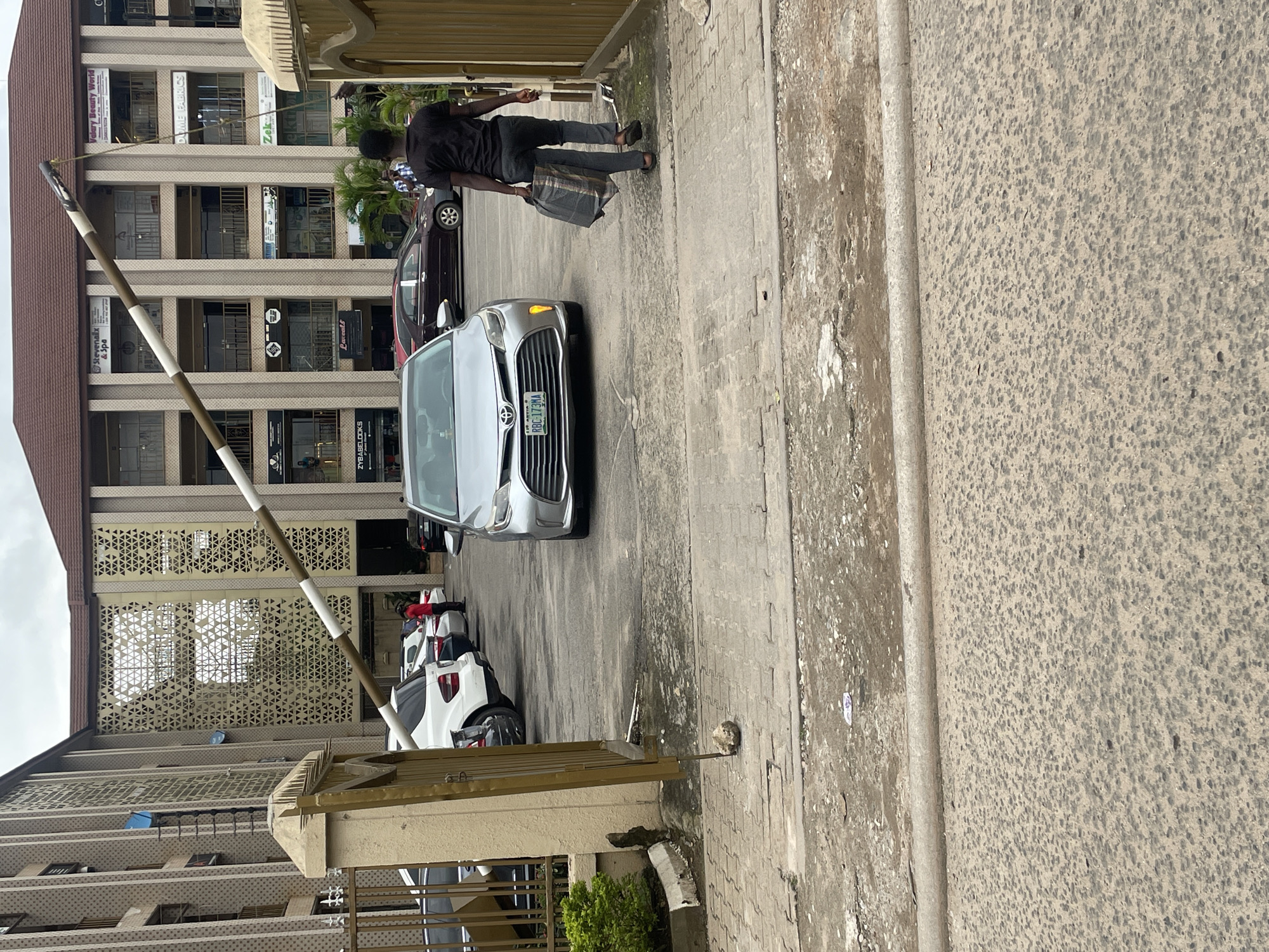 In Abuja, Nigeria, a silver Toyota minivan, with license plate RBC-173MA, is seen exiting a secured area through an open gate. A young man in a dark t-shirt and dark pants walks away from the camera, carrying a patterned bag in his right hand. He is walking to the right of the minivan. The gate is partially open, held up by a rock on the ground. Behind the vehicles, a multi-story building with shops on the ground floor is visible. The sky is overcast.