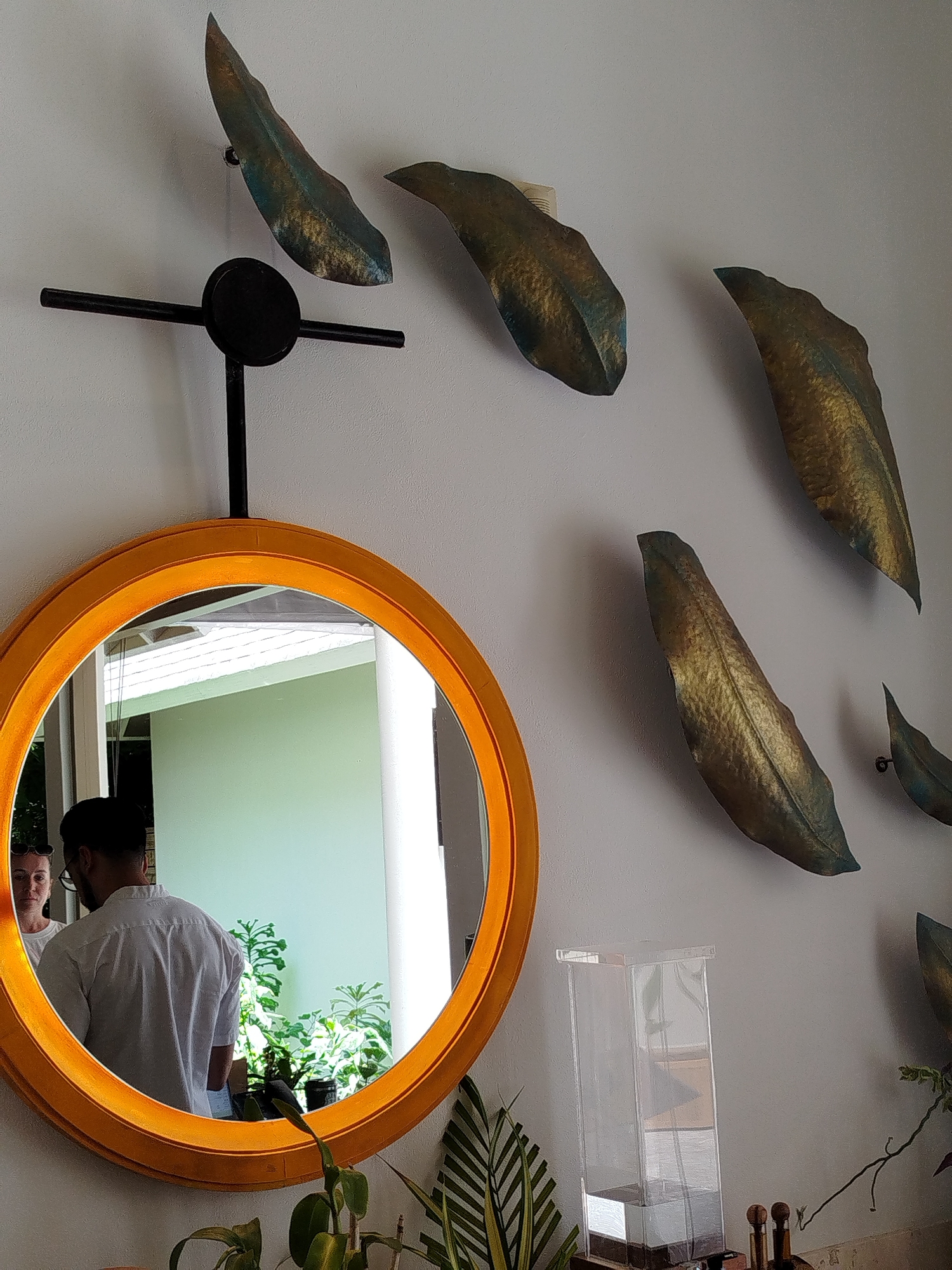 In a well-lit indoor setting, likely a home or hotel in Kendhoo, Maldives, an orange-rimmed circular mirror hangs on a pale wall adorned with decorative leaf-shaped art pieces. The mirror reflects a man wearing a white shirt and glasses, seen from behind, with a woman beside him. Beyond them, lush green plants are visible, suggesting an outdoor area with natural light. A clear acrylic container sits on a surface below the wall art, and further to the right, some wooden objects and a plant with striped leaves are present. The overall ambiance appears relaxed and peaceful. The time of day seems to be daytime, judging by the bright natural light illuminating the scene. There is no visible text in the image.