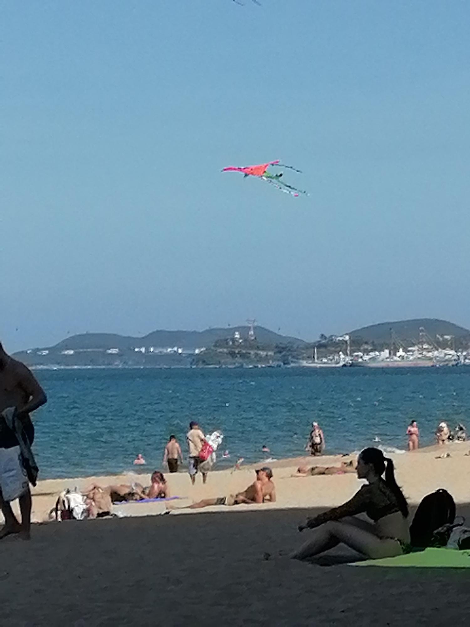 The scene depicts a beach on a sunny day. In the sky, a bright pink and green kite is flying. People are on the beach, some swimming in the ocean, others sunbathing or walking along the shore. A woman sits in the foreground on a light green towel, while a black backpack leans against her. Part of a man is visible on the left side. The background shows a coastline with hills. The location is in the city of Nha Trang Ward, Vietnam.