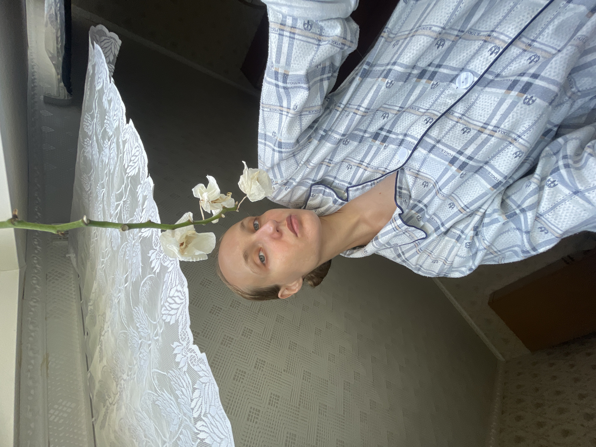 A young woman with light brown hair pulled back from her face looks directly at the camera in a close-up selfie. She is wearing light blue and white plaid pajamas with navy blue trim. A stem with three wilting white flowers hangs from above and to the left of her face. The background consists of a white lace curtain, a textured ceiling, and a dark wall. The scene appears to be indoors. The location in the city of Sharkan, Russia is not discernible from the image.