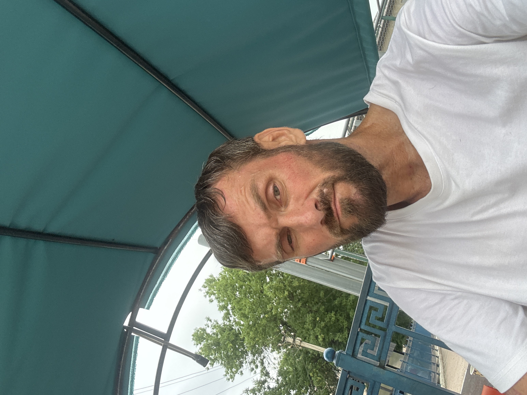 A middle-aged man with short brown hair and a beard, wearing a white t-shirt, is pictured from the chest up. He has reddish cheeks and is looking directly at the camera with a slightly squinted left eye. The man is seated outdoors, under a teal-colored awning supported by dark poles. In the background, there is a blue metal fence with a Greek key pattern and green trees. The sky is overcast.
