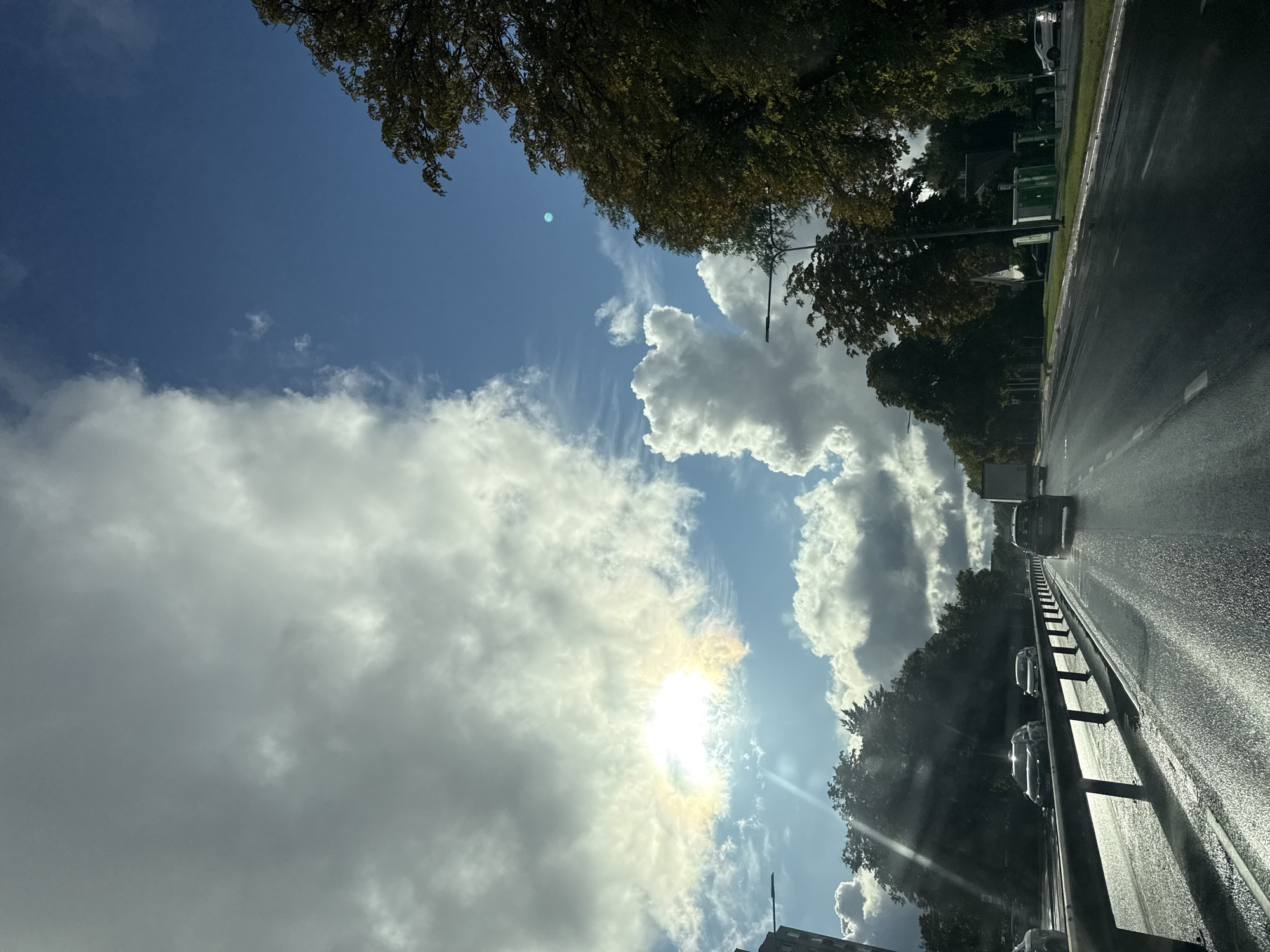 The image shows a road with cars traveling on it, viewed from the perspective of a car. The road appears to be wet, suggesting it might have recently rained or is still raining. Trees line the side of the road, and the sky is visible above, with a mix of clouds and patches of blue. The sun is partially obscured by clouds, casting a bright glow and some lens flare. The overall scene suggests a daytime drive on a road, possibly a highway or a main road in a suburban or rural area. There are no visible people, and the dominant objects are the cars and the surrounding environment. The presence of trees and the style of the road barriers might offer some clues about the specific location, which is noted as Tallinn, Estonia. The mood is neutral, reflecting a typical travel scene. No text is visible in the image.