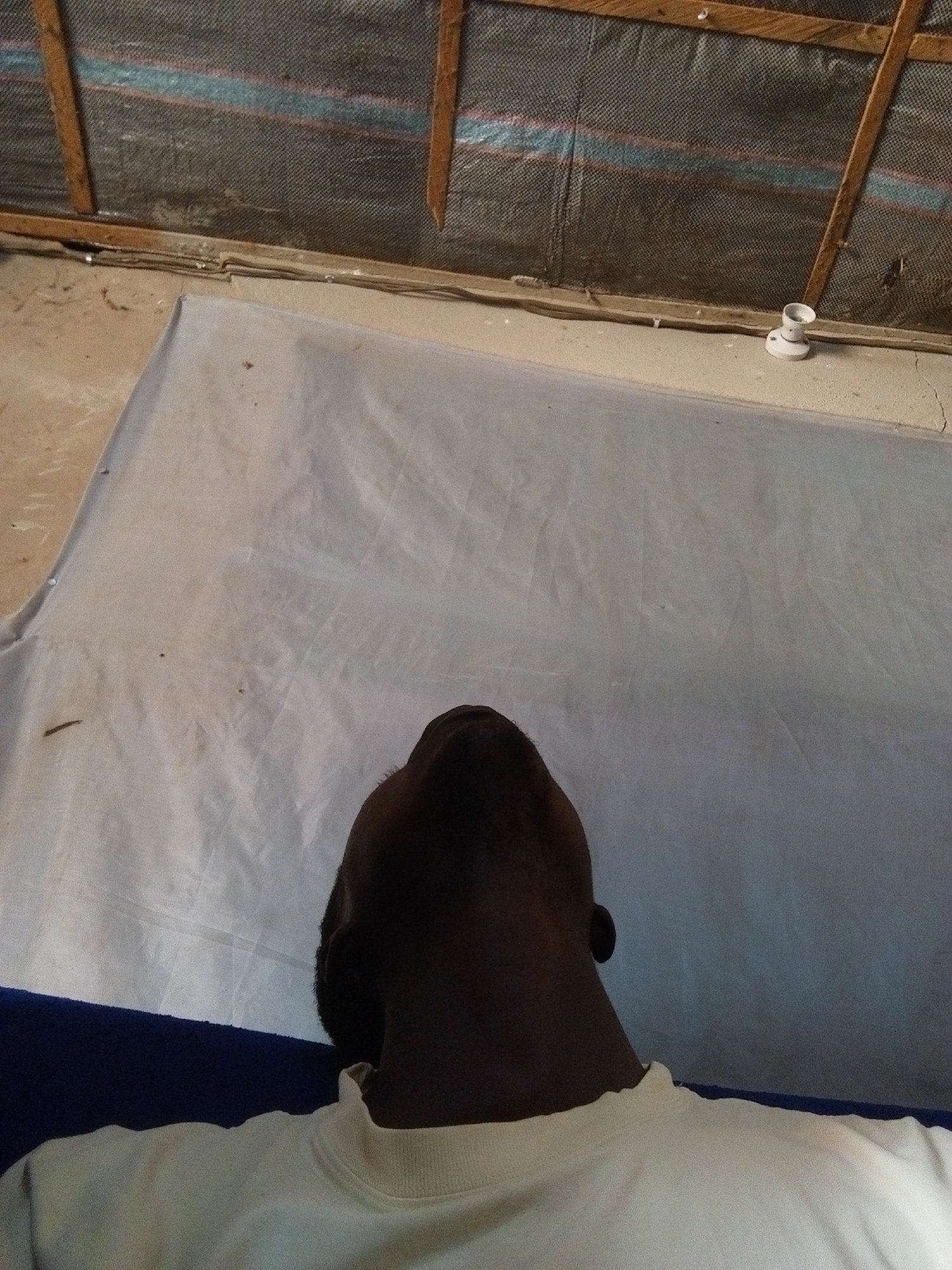 A person is lying on a white sheet, viewed from a high angle, looking upwards. Their head and neck are visible, with dark hair and skin. They are wearing a light-colored crew neck shirt. The white sheet is spread out on a floor that appears to be concrete. Above the sheet, there is a wooden structure supporting a dark mesh material, possibly a ceiling or wall covering. A small white object, resembling a light socket, is visible on a ledge in the background. The environment appears to be indoors, possibly in a sparsely furnished room in Nyabe, Nigeria.