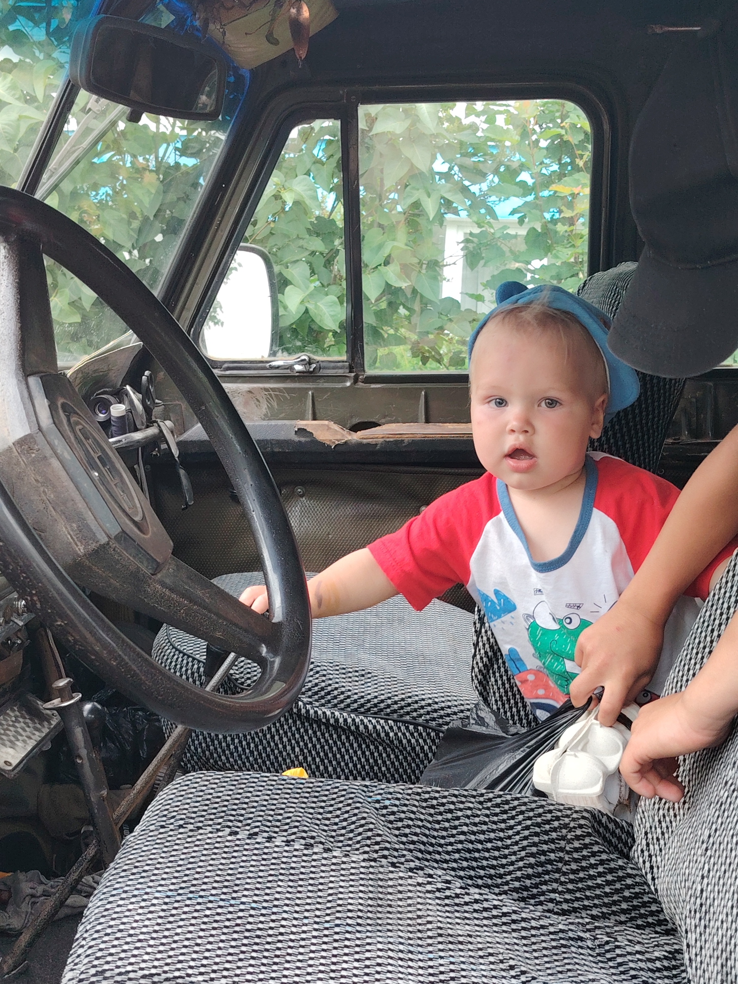 The image depicts the interior of a vehicle, possibly a truck, viewed from the passenger side. A young child, wearing a blue cap and a red and white t-shirt with a cartoon frog design, is seated in the driver's seat. The child's hands are visible, and they appear to be holding a small white object, possibly a toy or a container. To the right of the child, an adult's hands are also visible, reaching towards the child. The steering wheel is prominently positioned on the left side of the frame. The seats are covered with a black and white patterned fabric. The background visible through the windows consists of green foliage and a partially visible structure, suggesting an outdoor or semi-outdoor setting. The overall scene is casual and appears to be a candid moment. The image does not contain any specific visual cues that would indicate its location in Chaikovsky, Russia.