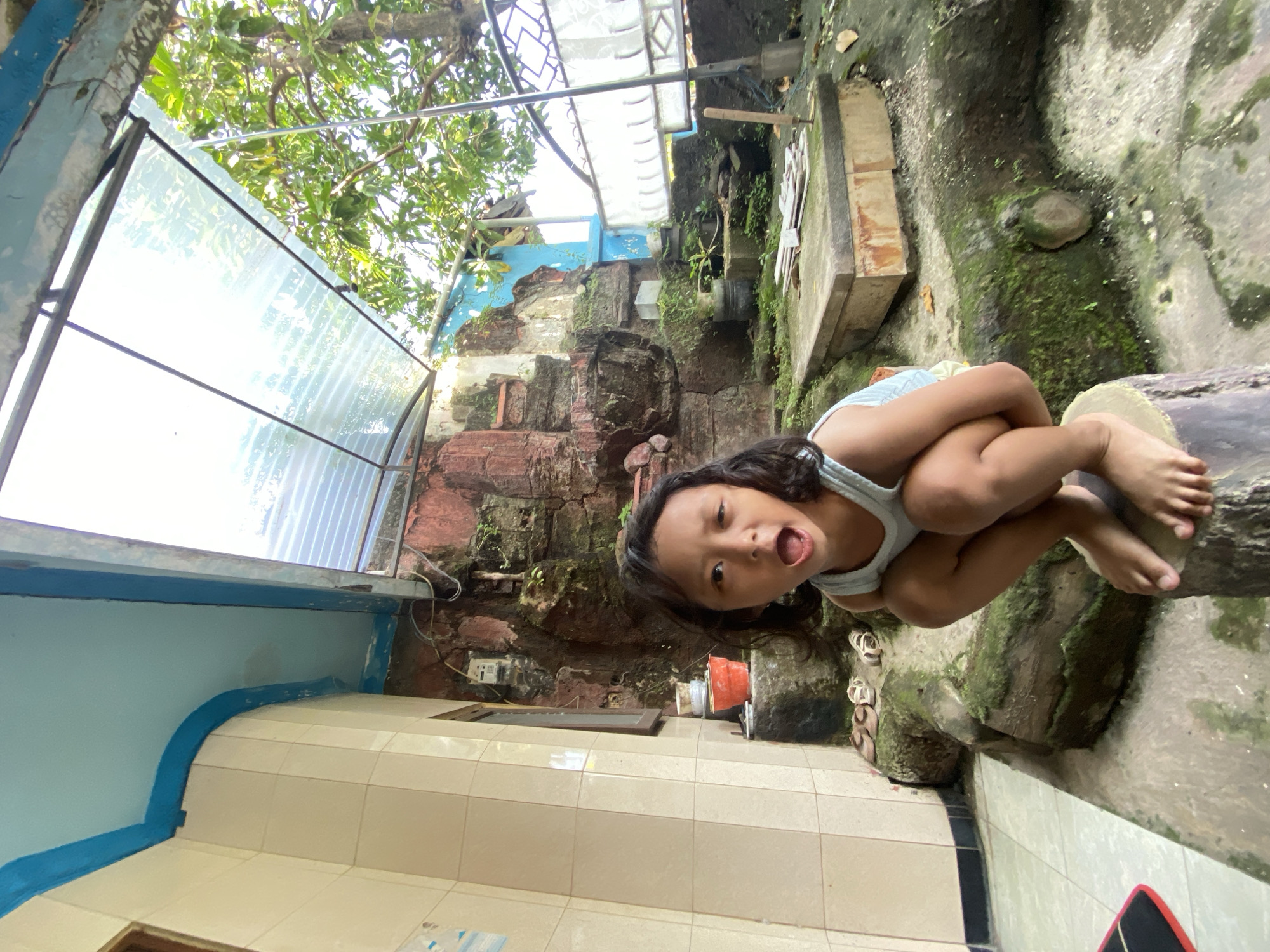 A young child is squatting on a mossy log in Gelam, Indonesia, captured in a tilted, medium shot. The child has long, dark hair and wears a light-colored tank top. They are looking directly at the camera with their mouth open. Behind the child, there is a wall composed of red bricks and cement structures. Moss and greenery are visible on the wall. To the left, a tiled wall and a clear, corrugated roof structure are present. The ground slopes downwards to the right.