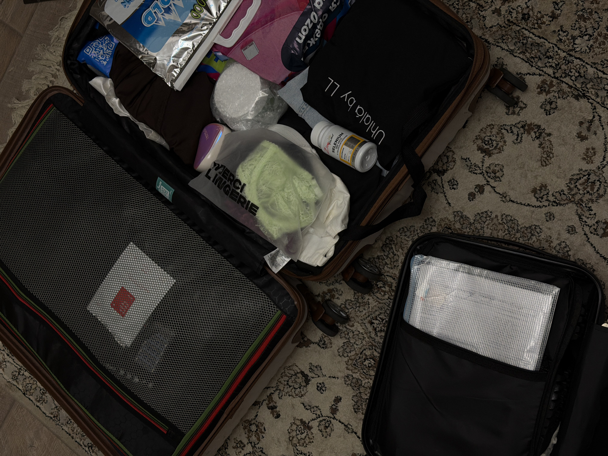 The image shows two suitcases partially packed and lying on a patterned rug. One suitcase is light brown with black trim and a mesh divider, revealing a small square white object with a red logo. The other suitcase is black and contains a clear, zippered pouch holding what appears to be papers. The open light brown suitcase contains various items including a bag labeled "COLD" and another bag labeled "ozon," a roll of white material, a bottle of "SELENIUM" supplement, and a package labeled "MERCI LINGERIE." A black bag labeled "Uhlala by LL" is also visible. The scene suggests someone is packing or unpacking their luggage. The location, Nakhodka, Russia, is not discernible from the image.