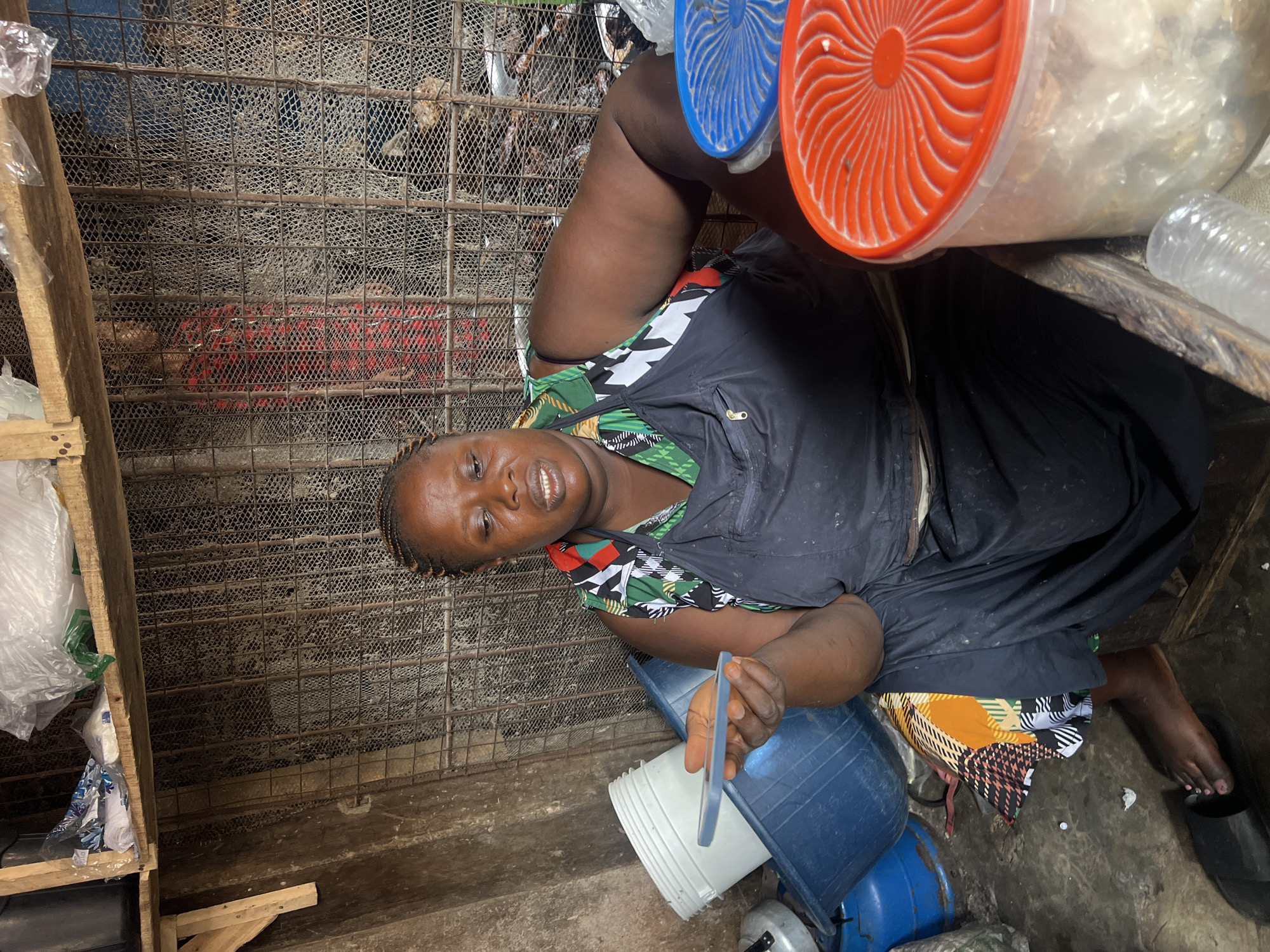 A dark-skinned woman with braided hair and an apron sits in what appears to be a market stall in Nkpor, Nigeria. She is holding a mobile phone in her right hand. She wears a green and white patterned top under a black apron. Behind her, a metal grate fence separates her from what looks like more goods. To her left are several plastic containers and buckets, one white and one blue, filled with items. To her right, a large clear plastic container with an orange lid and another with a blue lid are visible. The overall environment appears to be an indoor market stall with concrete walls and wooden shelving above. The lighting is dim.