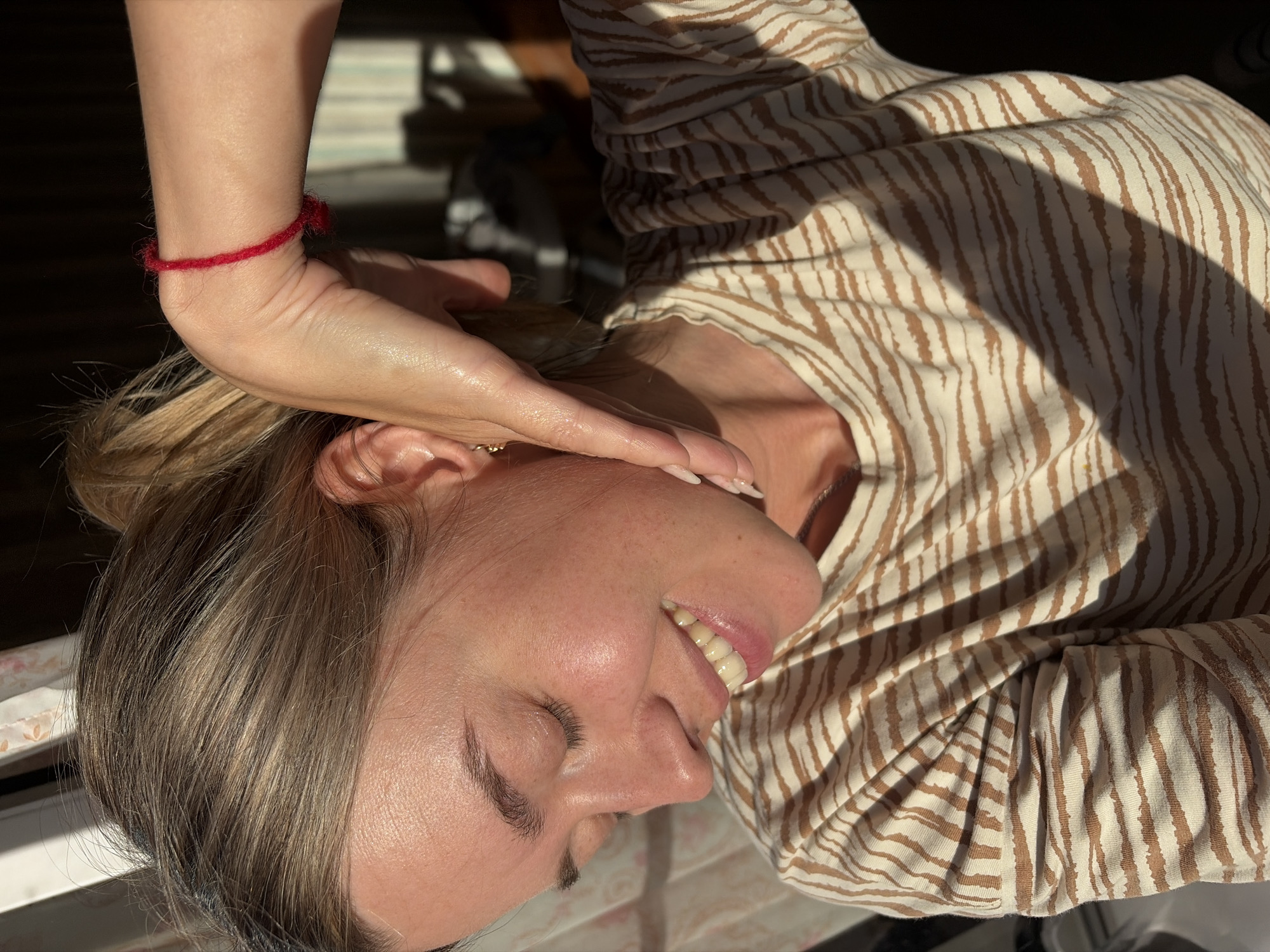 A close-up, side-profile shot shows a woman's face and arm. She has blonde hair pulled back, and her eyes are closed, with a slight smile on her lips. Sunlight catches her skin, highlighting its texture and a scattering of freckles. She is wearing a light-colored top with a brown zebra-like stripe pattern. A red string bracelet is visible on her wrist. Her arm is bent, and a hand, with manicured nails, gently rests on her forehead, partially obscuring her eye. The lighting suggests it's daytime, with strong shadows cast on her clothing and in the background, hinting at sunlight streaming through blinds or a window. The background is softly blurred, with dark areas and lighter patches that could indicate furniture or a room interior. The overall mood is peaceful and relaxed, perhaps indicating a moment of repose or self-care.