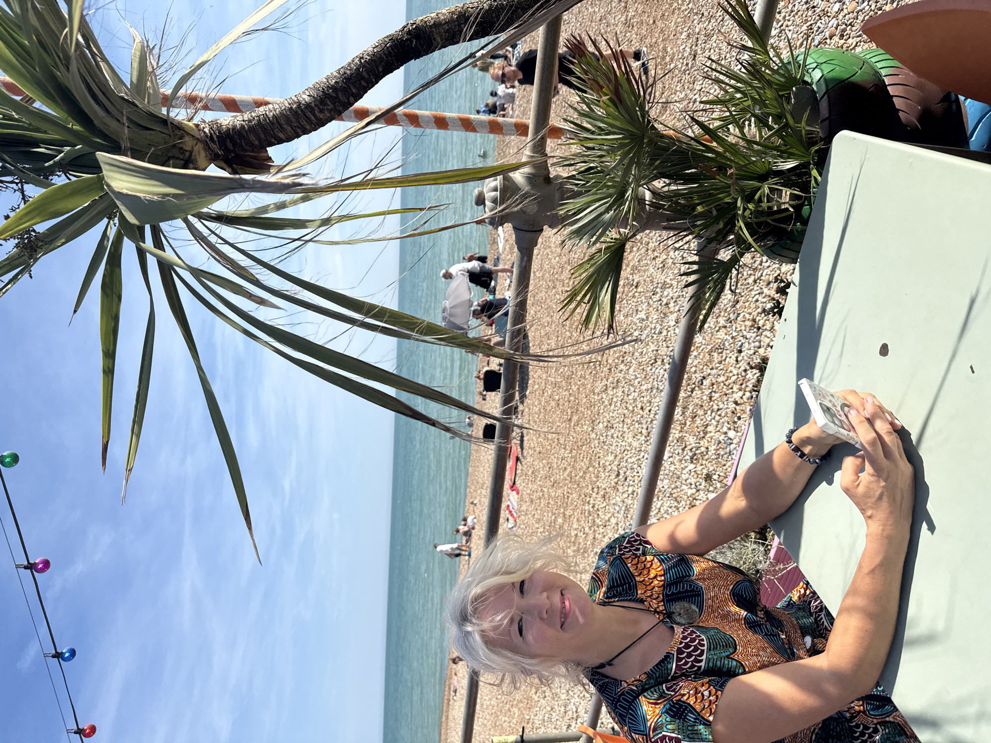 A woman with blonde, wavy hair is seated at a table outdoors with a smartphone in her hands. She is wearing a vibrantly patterned dress with orange, blue, and green accents. In the background, a beach is visible with people scattered across the pebbles and in the turquoise sea. A palm tree with a striped trunk grows nearby, and string lights with colored bulbs are strung across the sky. The scene suggests a sunny day at a coastal location in St Leonards, United Kingdom.