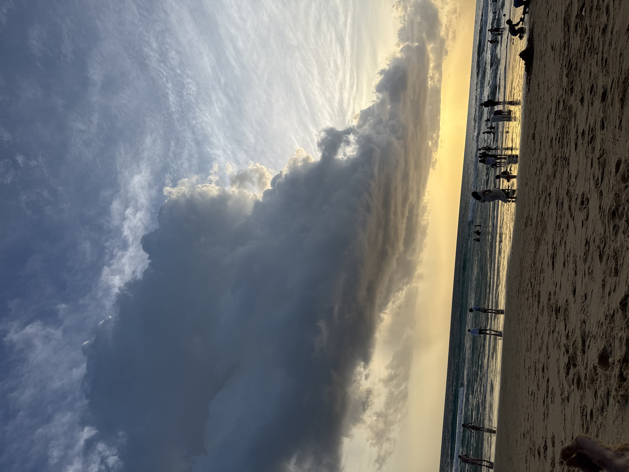 The image depicts a sunset scene at Karon Beach, Thailand. The sky is filled with dramatic clouds, with a large, dark storm cloud dominating the left side, contrasting with a bright, yellow sunset glow on the horizon. Wispy white clouds stretch across the upper right portion of the sky. The ocean is visible in the midground, with gentle waves rolling onto the sandy shore. Several people are scattered along the waterline and in the shallow water, some wading and others standing. In the foreground, the sandy beach is in shadow, with the feet of the photographer visible in the bottom left corner. The overall atmosphere suggests a transition from day to evening, with a hint of impending weather.