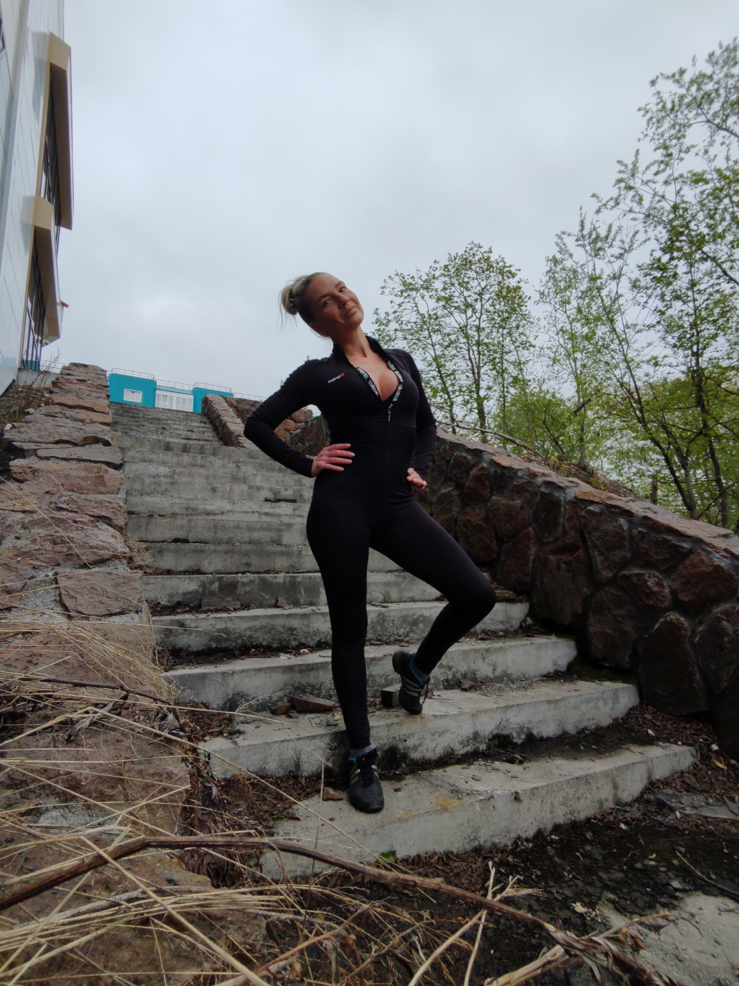 A woman wearing a black, form-fitting jumpsuit with a zippered neckline is standing on a concrete staircase. The staircase is bordered by a low stone wall and surrounded by dry grass and sparse trees under an overcast sky. In the background, a modern building with a light-colored facade and blue accents is visible. The woman has her hands on her hips, with her head tilted back and to the left. The overall scene suggests an outdoor urban setting in Petropavlovsk-Kamchatsky, Russia, possibly a park or recreational area with a view of a building.