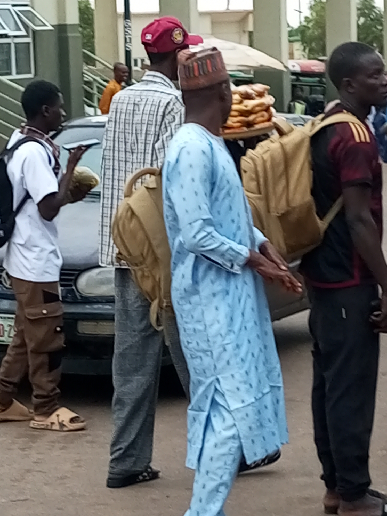 The image shows a group of people in what appears to be an outdoor market or roadside setting in Gombe, Nigeria. In the foreground, a man wearing a light blue traditional outfit with a patterned design and a cap is standing. He is holding his hands together in front of him. To his right, another man, partially visible, is wearing a dark red shirt with yellow stripes on the sleeves and a backpack. He is facing away from the camera. Further to the left, a man in a white short-sleeved shirt and brown cargo pants is holding a round object, possibly food. He is wearing sandals. Behind him, a car is visible, with a license plate that appears to start with "GOM." The background shows a building with windows and a covered stall, suggesting a commercial area. The ground is paved, and there are other people in the background, including one wearing an orange shirt. The lighting suggests it is daytime, possibly overcast. The overall scene depicts a busy, casual street environment.
