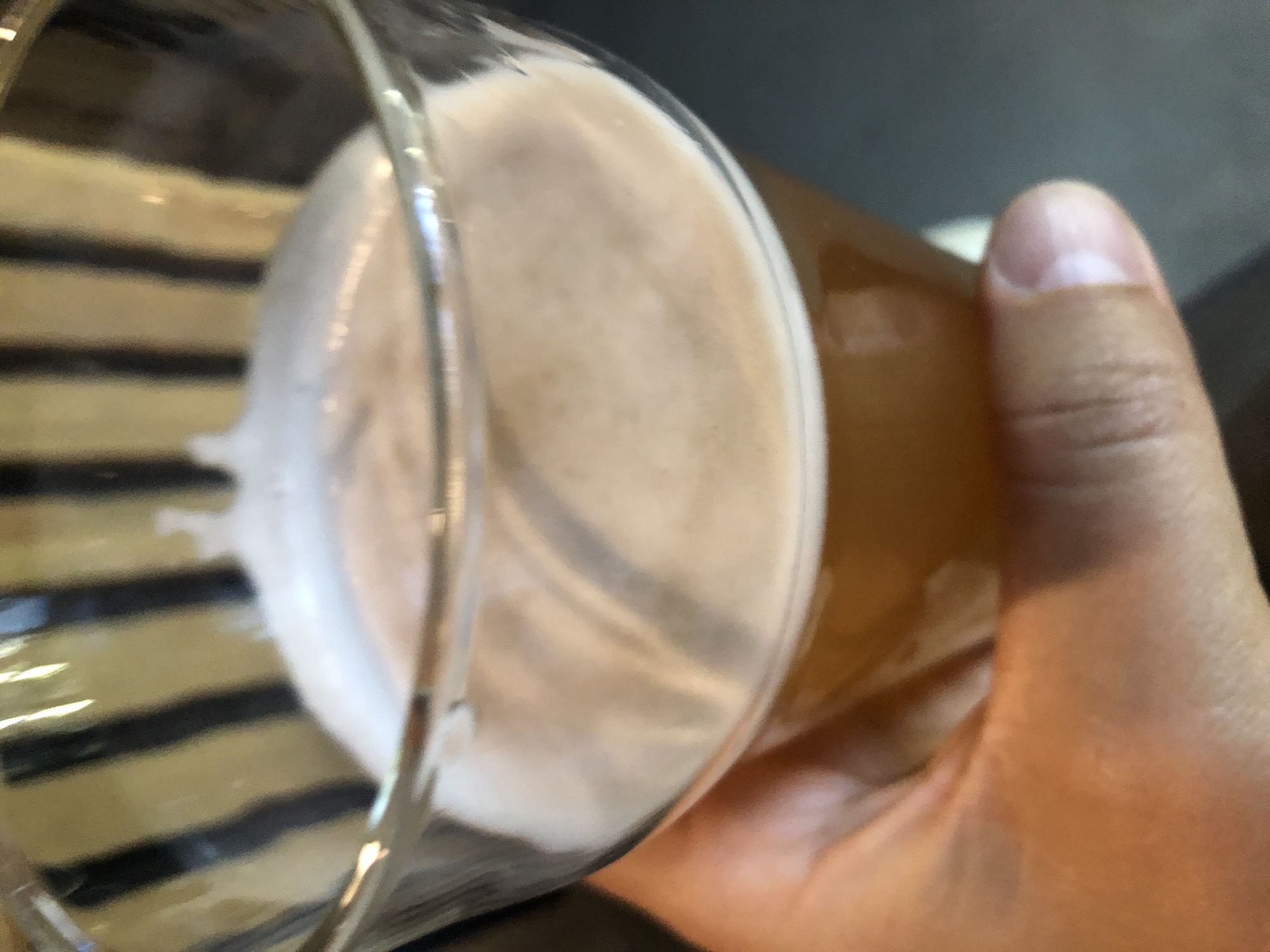 A close-up shot of a glass of beer, held by a person's hand, with a blurry background of vertical stripes. The beer has a foamy head and is a golden-amber color. The person's thumb and part of their fingers are visible, gripping the base of the glass. The background appears to be a dark, out-of-focus surface with a pattern of dark vertical lines on a lighter background. No specific location in Saint Petersburg, Russia, can be determined from the image.