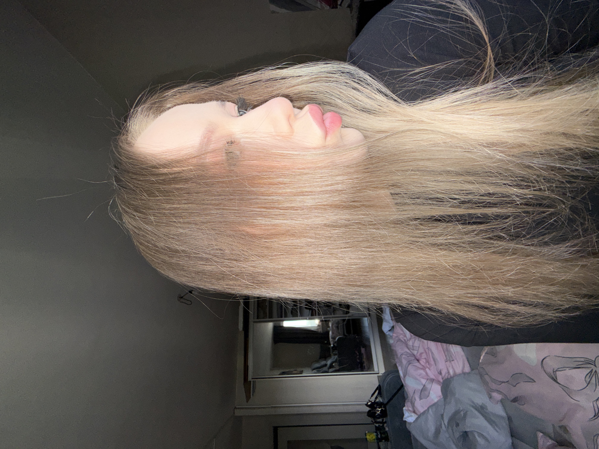 The image displays a close-up, profile shot of a young individual, likely female, positioned indoors. The subject's long, straight, light blonde hair falls forward, partially obscuring the face, with several individual strands visibly uplifted. The face is turned towards the right side of the frame, revealing fair skin, prominent eyelashes (suggesting cosmetic enhancement), and a lightly defined eyebrow. The lips are full, pink, and conspicuously pursed. A black long-sleeved garment is worn.
To the immediate left of the subject, a grey pillow and a pink patterned blanket or duvet are visible on what appears to be a bed or sofa. The background is dominated by a large, mirrored wardrobe or closet, which reflects the room beyond. The reflection reveals an interior space with a window (serving as a light source) fitted with dark curtains, a chair, and what appears to be another bed or sofa. Above the mirrored unit, an exposed wooden or structural element is visible. To the far left of the frame, a doorway is partially discernible, along with small objects (bottles, a dark bag) on a nearby surface. The overall lighting is somewhat dim, suggesting natural light from the reflected window as the primary source. The specific urban location of Chaikovsky, Russia, cannot be visually confirmed from the details present within the image.