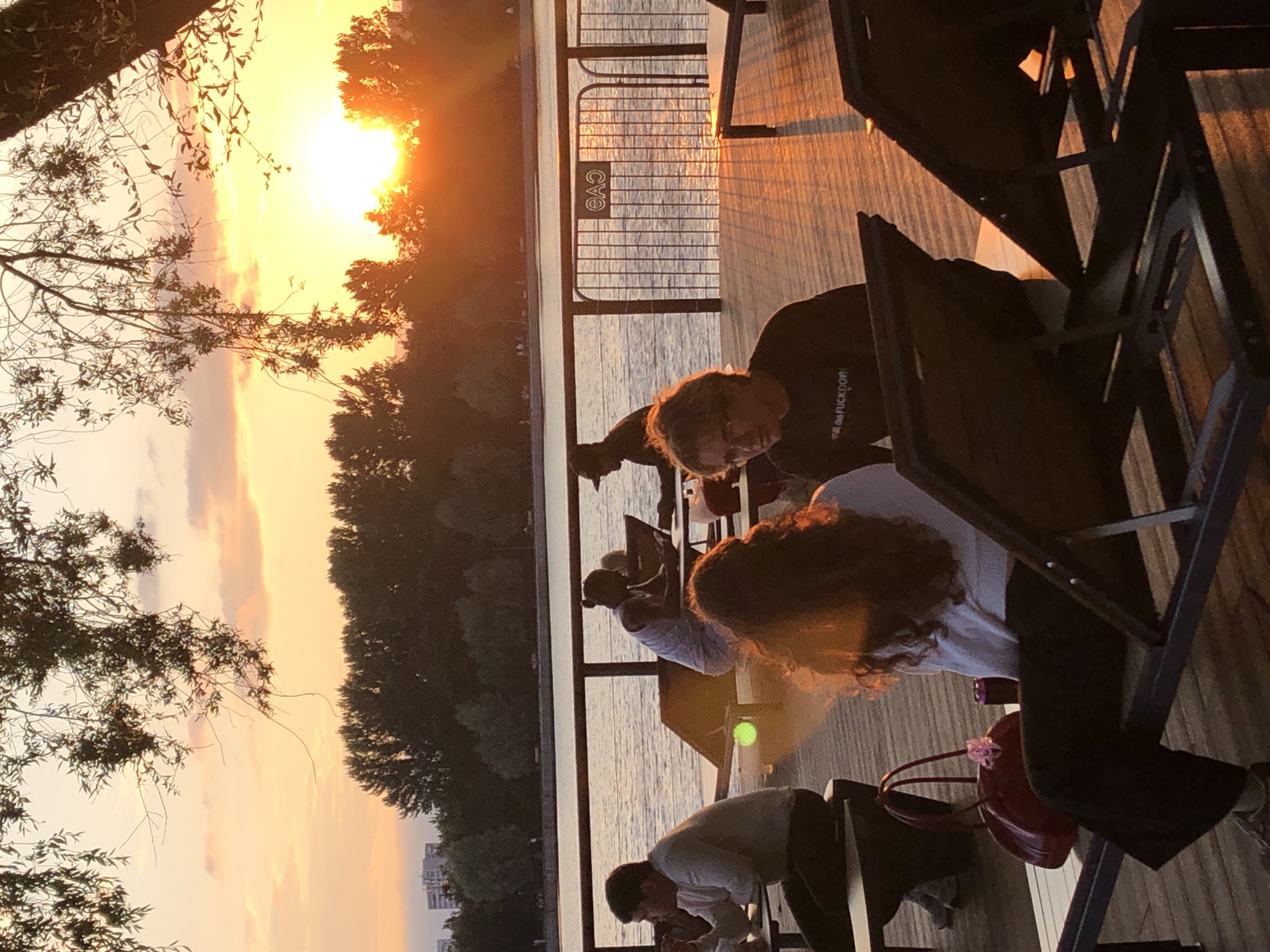 The image depicts an outdoor seating area next to a body of water during sunset. Several people are visible, some seated at tables and others relaxing on lounge chairs. The sky is a warm orange and yellow hue, with trees silhouetted against it along the far bank of the water. A railing with a metal fence separates the seating area from the water. The overall impression is one of a relaxed evening gathering at a waterfront establishment. The scene is likely a restaurant or cafe with outdoor seating, potentially located in Khimki, Russia, given the context of the request.