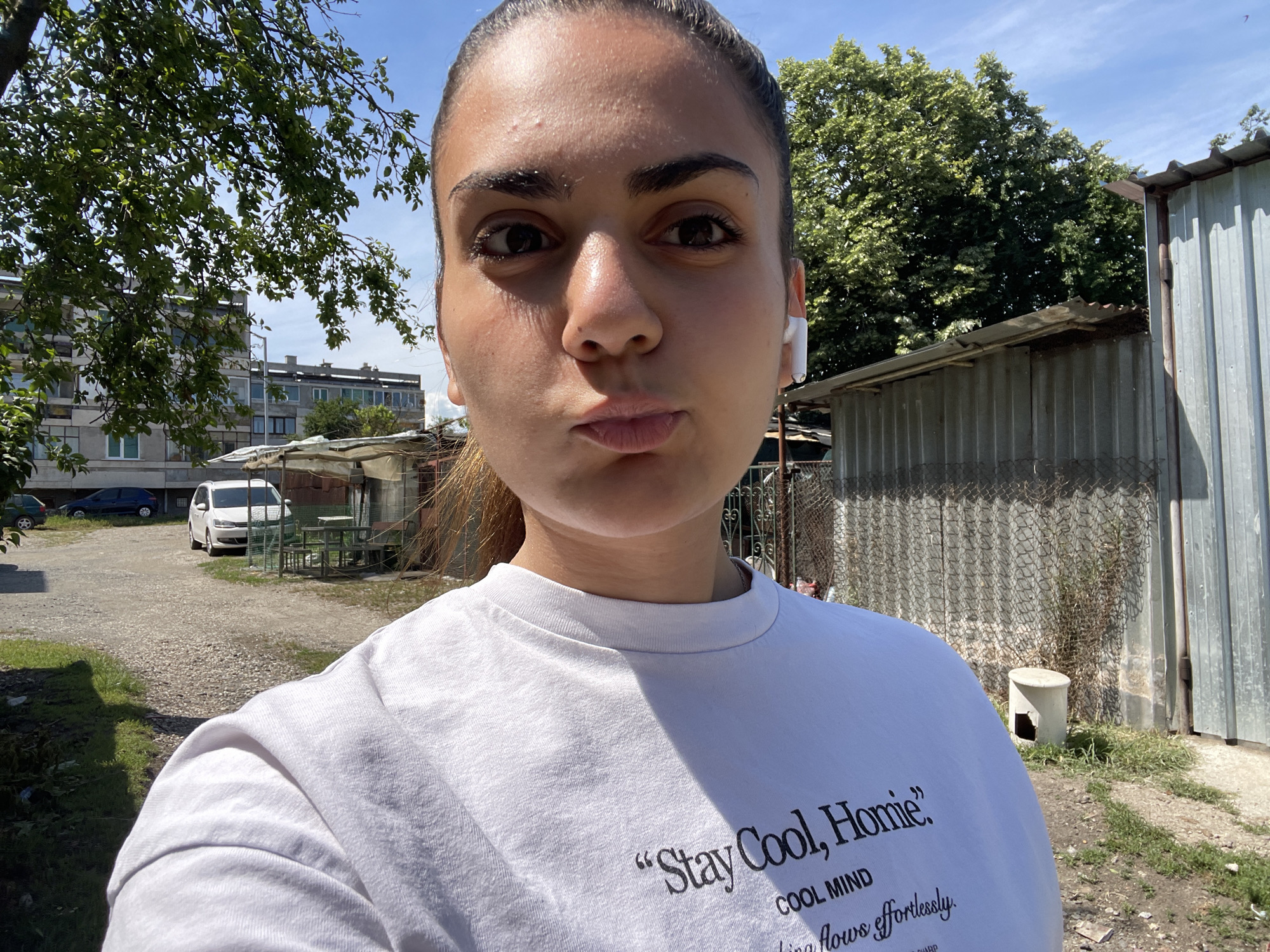 A young woman is taking a selfie outdoors in what appears to be an urban or semi-urban area. She is wearing a white t-shirt with text on it, and has earbuds in. She is looking directly at the camera with her lips pursed. In the background, there are some trees, a building, a parked car, and some corrugated metal structures. Based on the context provided, the scene is located in the city of Debelt, Bulgaria.