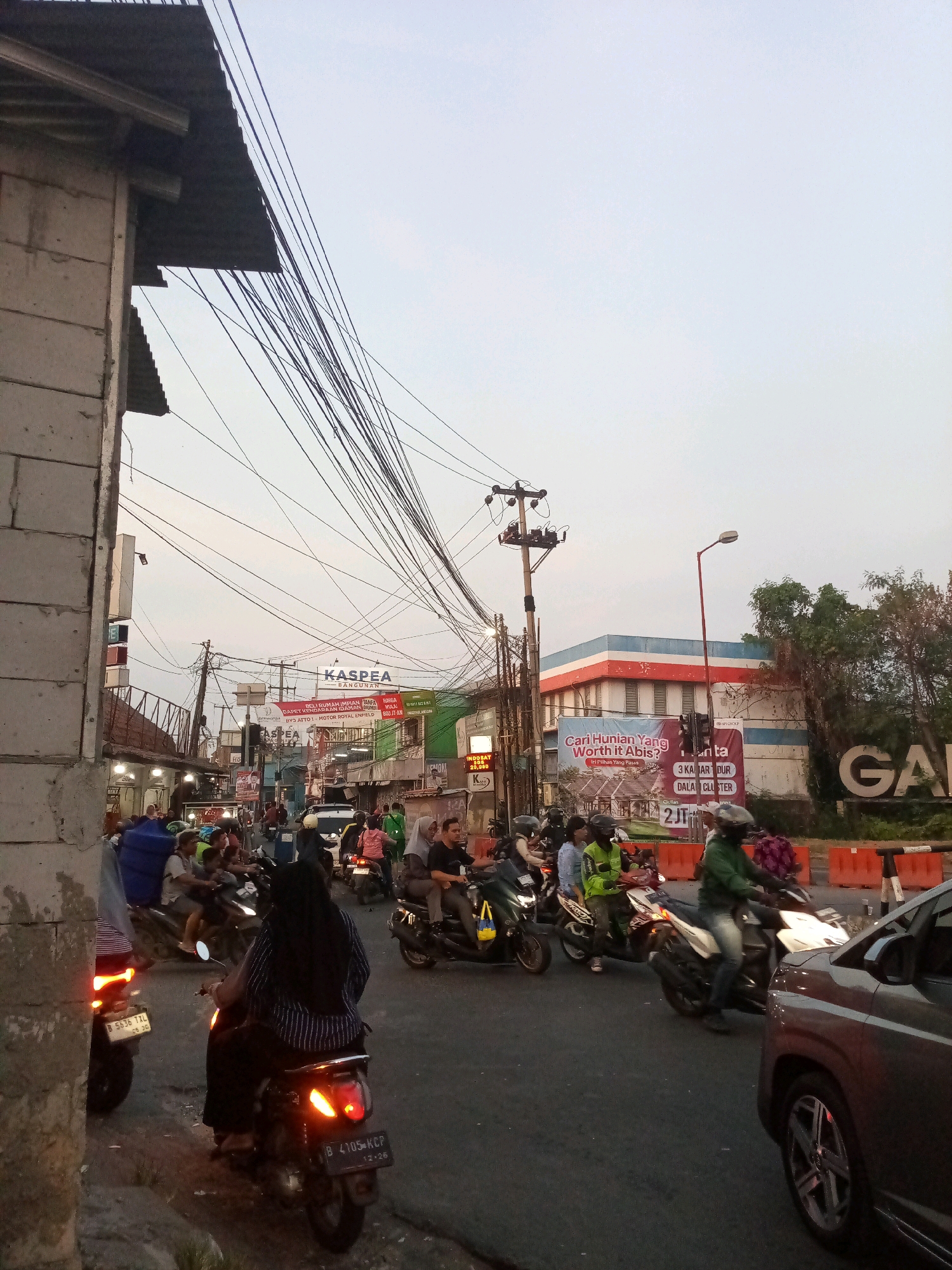 The image captures a street scene in Karangsatria, Indonesia, characterized by a moderate amount of traffic, primarily motorcycles. The sky is overcast, suggesting it might be late afternoon or early evening. The road is paved, and there are buildings lining both sides of the street, some of which appear to be commercial establishments with signage. Overhead, a dense network of electrical wires crisscrosses the sky, a common sight in urban areas of Indonesia.
On the right side of the road, a prominent billboard advertises housing with the text "Cari Hunian Yang Worth It Abis?" (Looking for Housing That's Worth It?). Below this, smaller text mentions "3 KAMAR DARI DALAM CLUSTER" (3 bedrooms from within the cluster) and "2JT-AN" (starting from 2 million). Further to the right, a large sign with the word "GANDA" is visible. A building with a red and white facade is also present in the background.
Numerous individuals are depicted on motorcycles, some in motion and others seemingly stopped in traffic. The riders are dressed in everyday attire, with some wearing helmets. One woman in the foreground is riding a scooter, her dark hijab and striped shirt clearly visible. Another motorcycle in the foreground has a license plate that reads "B 4105 KCP". To the left, a partial view of a brick building and a corrugated metal roof is visible, framing the left edge of the image. The overall impression is of a busy, ordinary day in a populated area of Karangsatria.