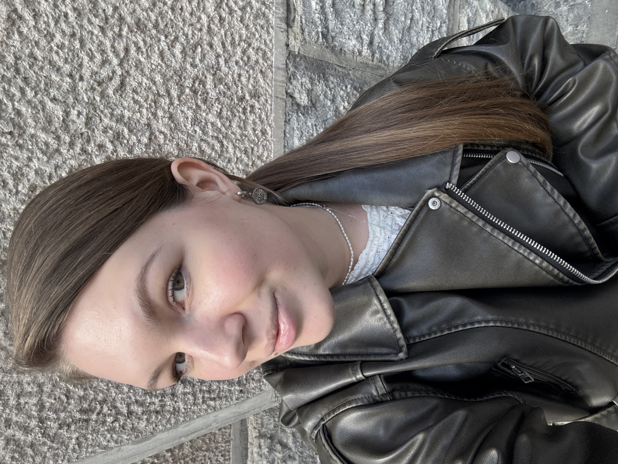 A close-up, slightly tilted portrait captures a young woman outdoors in what appears to be Belgrade, Serbia. She has light skin, long straight brown hair parted on one side, and light-colored eyes. Her gaze is directed calmly towards the viewer, with a subtle, neutral expression.
The woman is dressed in a black, leather-like biker jacket featuring multiple zippers and silver snaps, and a pointed collar. Beneath the open jacket, a white lace or intricately patterned top is visible. She wears a delicate silver chain necklace and a small, circular silver earring with an embossed design, possibly a compass rose, in her visible ear.
The background consists of a rough, textured grey stone or concrete surface, characteristic of an urban or architectural setting. The bright, natural light suggests it is daytime, likely a sunny day given the clear highlights and shadows on her face and clothing. The image primarily focuses on the woman's head and upper torso, without depicting any specific activity beyond posing for the photograph. There is no visible text in the image.