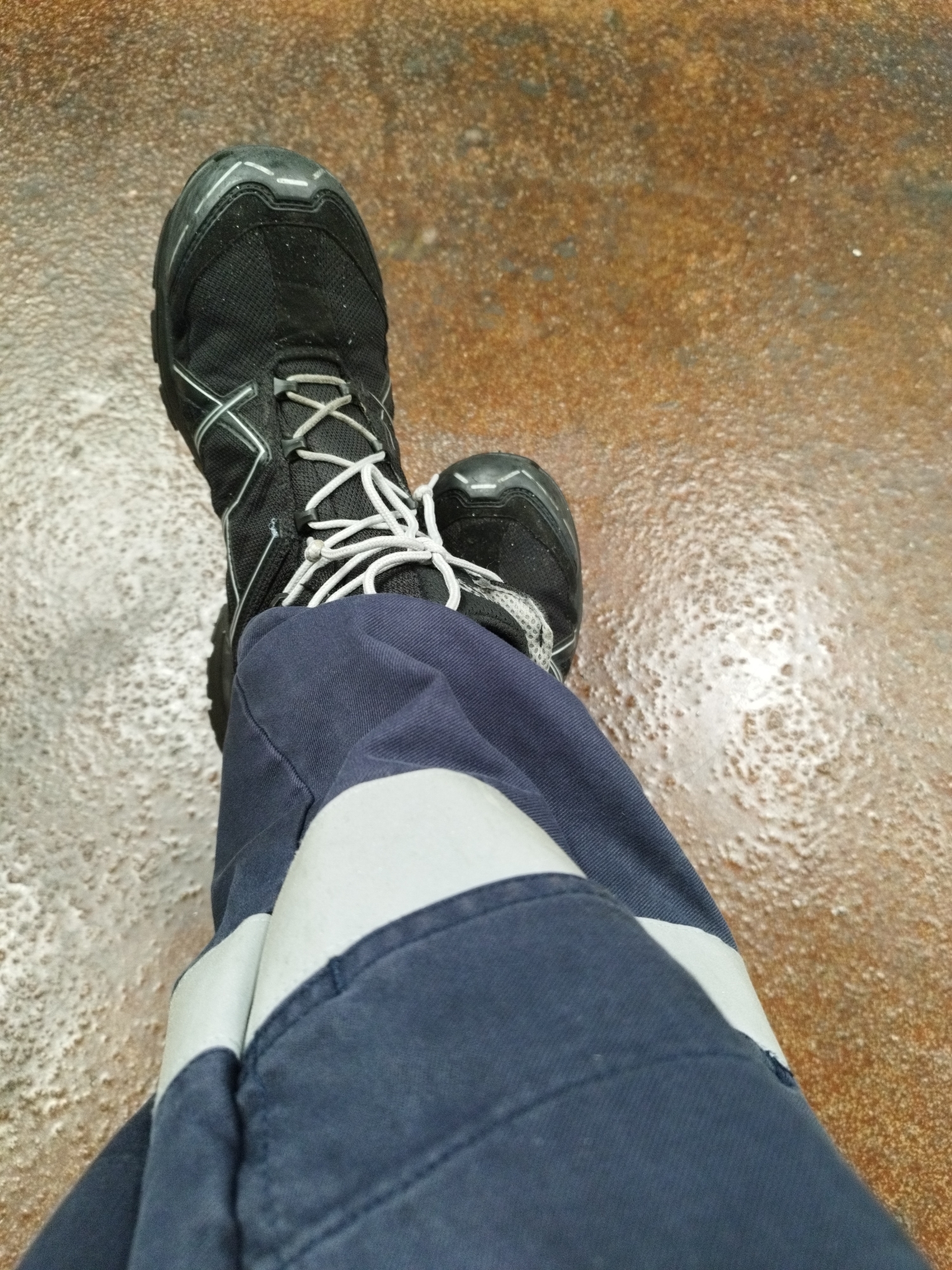 The image shows a close-up view of a person's legs and feet wearing dark blue work pants with reflective stripes and black hiking boots. The boots have white laces and grey accents. The person is standing on a wet, textured surface that appears to be concrete or asphalt, with areas of standing water and what might be salt deposits. The background is blurred, suggesting a shallow depth of field. The scene is likely outdoors, possibly in an industrial or port area, given the practical nature of the clothing and the condition of the ground. The lighting appears to be overcast or diffused. The overall impression is one of a person in work attire, in an environment that is damp and potentially gritty.