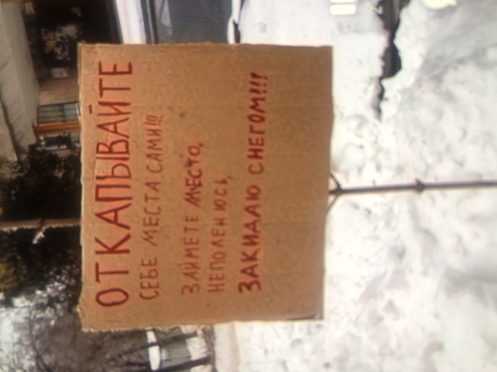 A cardboard sign on a pole reads in Russian: "CLEAR SPOTS FOR YOURSELVES. TAKE YOUR PLACE YOURSELF. I'M BURYING IT WITH SNOW!!!" The sign is in a snowy urban environment.