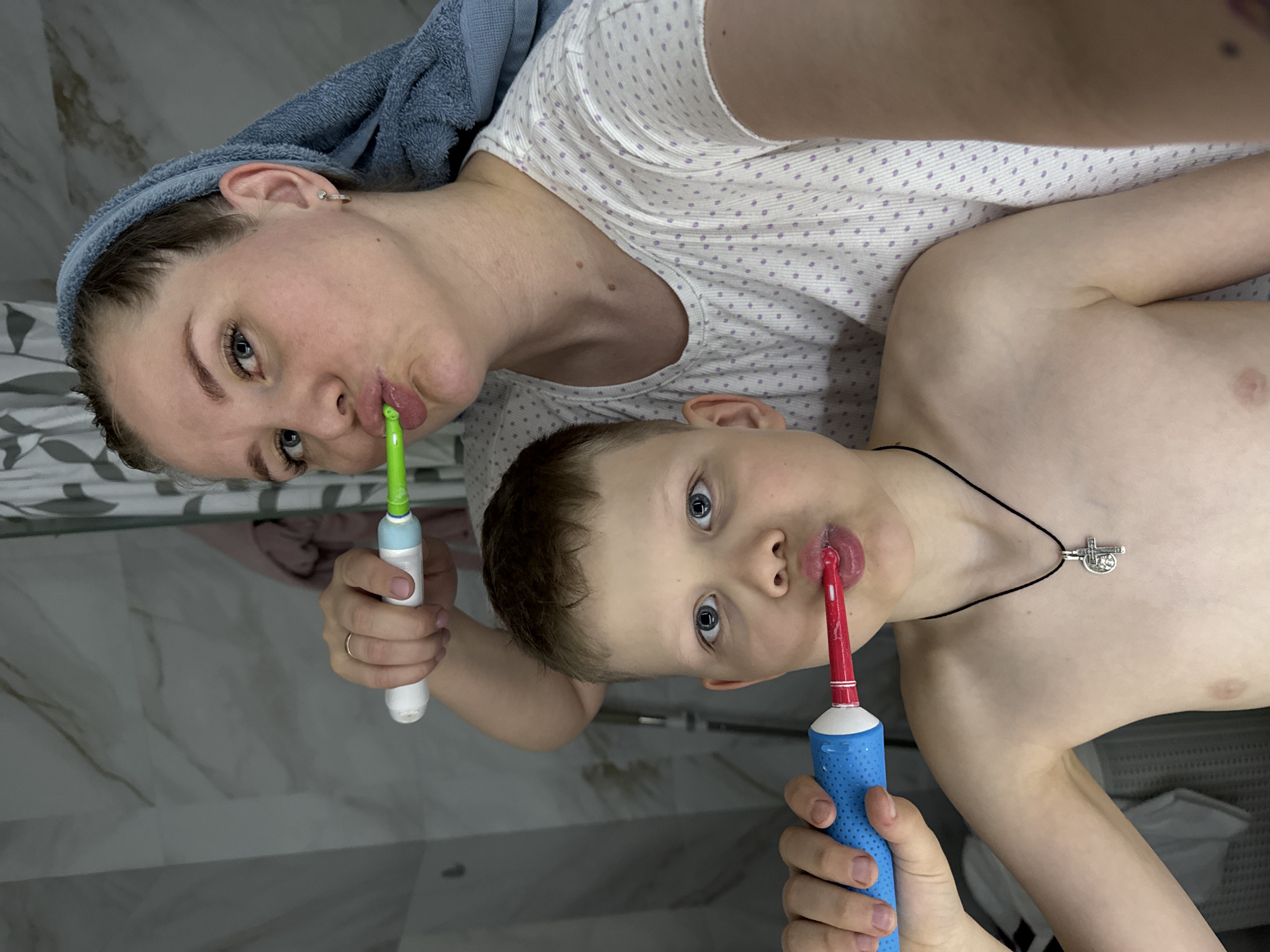 In a bathroom setting, a woman and a child are captured in a selfie while brushing their teeth. The woman, with a blue towel wrapped around her head, is holding a white and green electric toothbrush. The child is shirtless with a silver cross necklace, holding a blue and red electric toothbrush. The bathroom features tiled walls. This image appears to be taken in the city of Gornyi (Dvurechenskoe S/P), Russia.