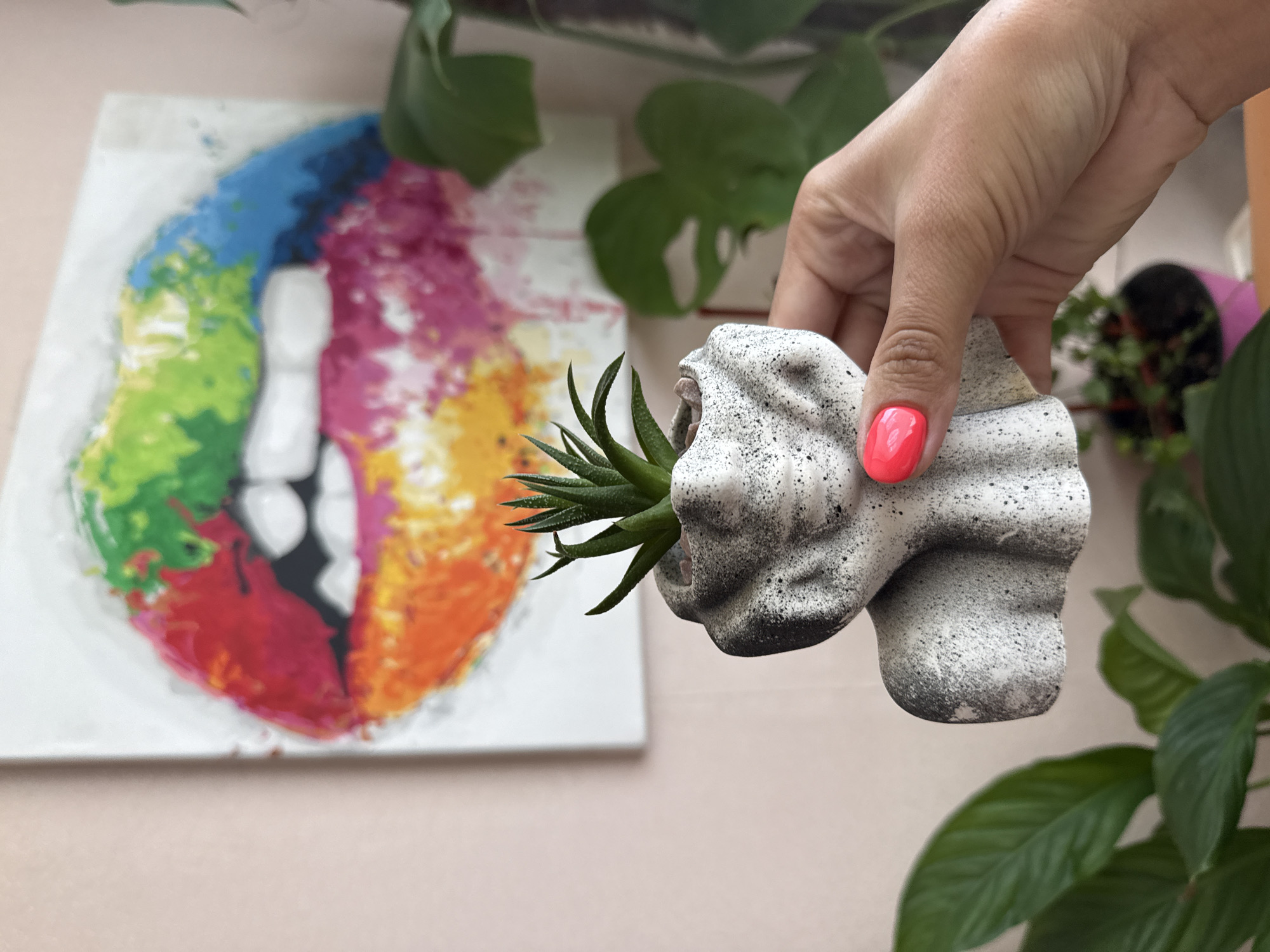A woman's hand with bright pink nail polish holds a small, concrete planter shaped like a bust with a succulent plant growing from its head. In the background, a painting of colorful lips is displayed on a pink wall. Green plants are visible in the foreground and background. The scene appears to be indoors.
