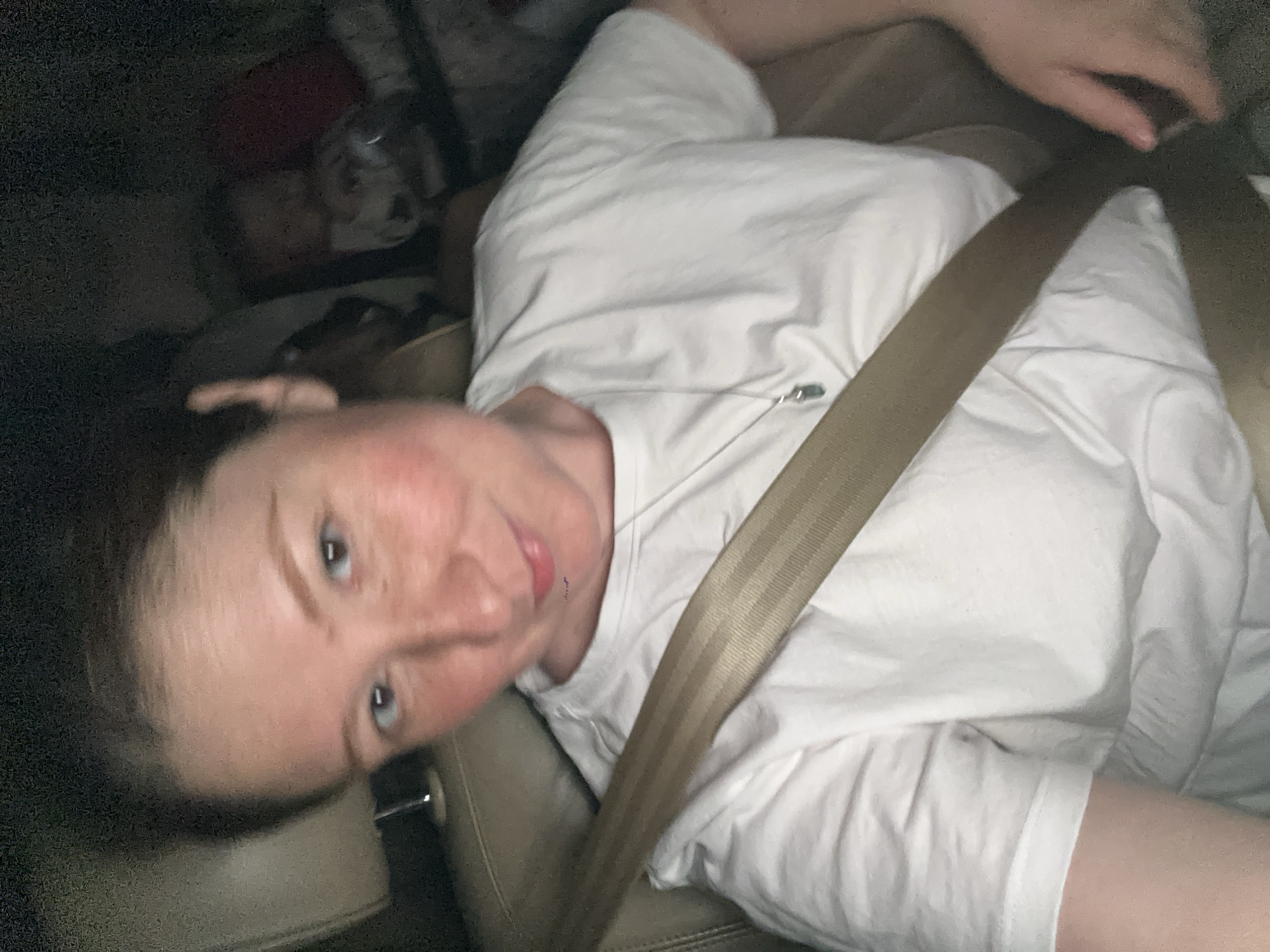 A person wearing a white t-shirt and secured with a beige seatbelt is visible in a car. The person has fair skin, blue eyes, and dark hair. The background is dark and indistinct. There are no visible landmarks to suggest the location is Nizhny Novgorod, Russia.