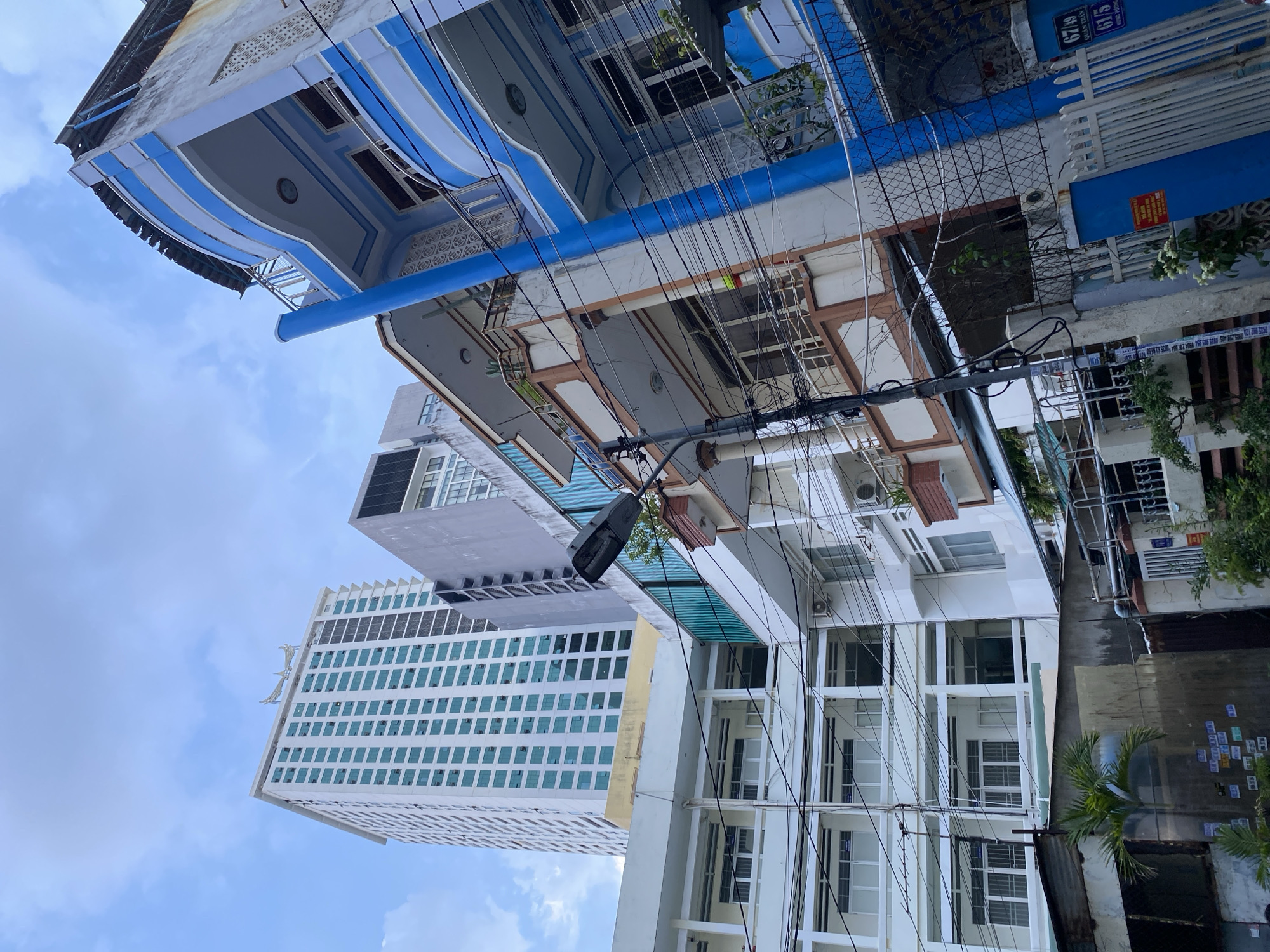 This media file captures an upward-angle view of buildings in Nha Trang Ward, Vietnam, under a cloudy sky. The scene is dominated by a variety of architectural styles, from older, ornate structures with blue and white accents to a modern, tall building with a facade of numerous rectangular windows. A dense network of overhead wires crisscrosses the frame, suggesting an urban environment with extensive electrical infrastructure.
The older buildings show intricate decorative elements, including patterned friezes and rounded balconies. The modern building, in contrast, is sleek and geometric, with its windows reflecting the cloudy sky. A street lamp is visible attached to a utility pole. The lighting suggests it is daytime, likely overcast, as there are no strong shadows. The overall impression is one of urban density and a mix of old and new construction.
Visible text includes street numbers on blue signs: "67/49" and "61/5". There is also a red sign on one of the older buildings that appears to be a warning or informational notice. Some smaller, square stickers with text are visible on a dark corrugated metal surface on the right side of the frame, but the text is too small to read. No people or specific activities are clearly depicted; the focus is on the urban landscape and its structures.
