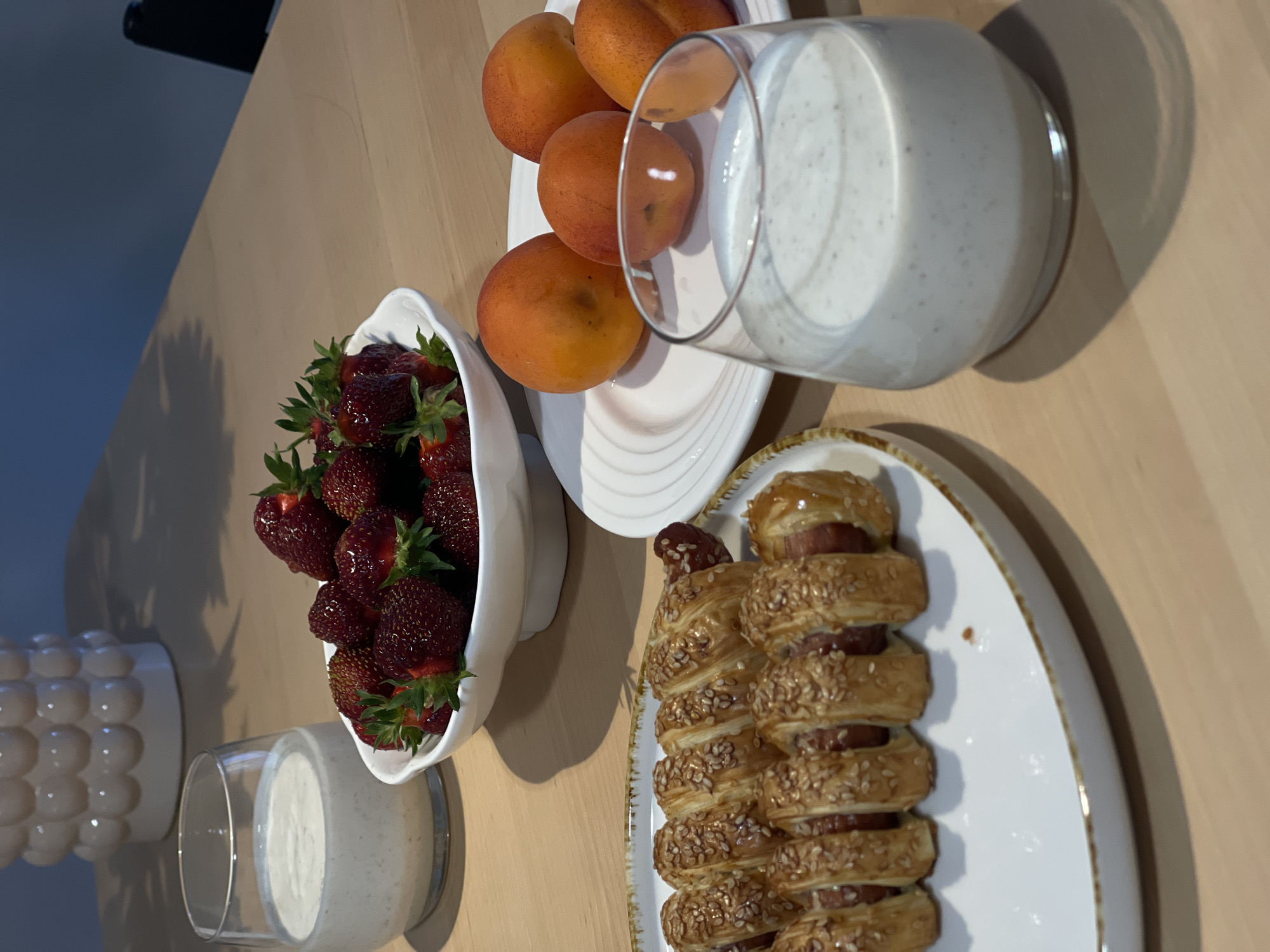 The image shows a wooden table with a meal. On the table, there is a white bowl filled with strawberries, a white plate with apricots, and a white plate with two sausage rolls. There are also two glasses containing a creamy white liquid. In the background, a textured white vase is visible. The scene appears to be indoors, possibly in a home.