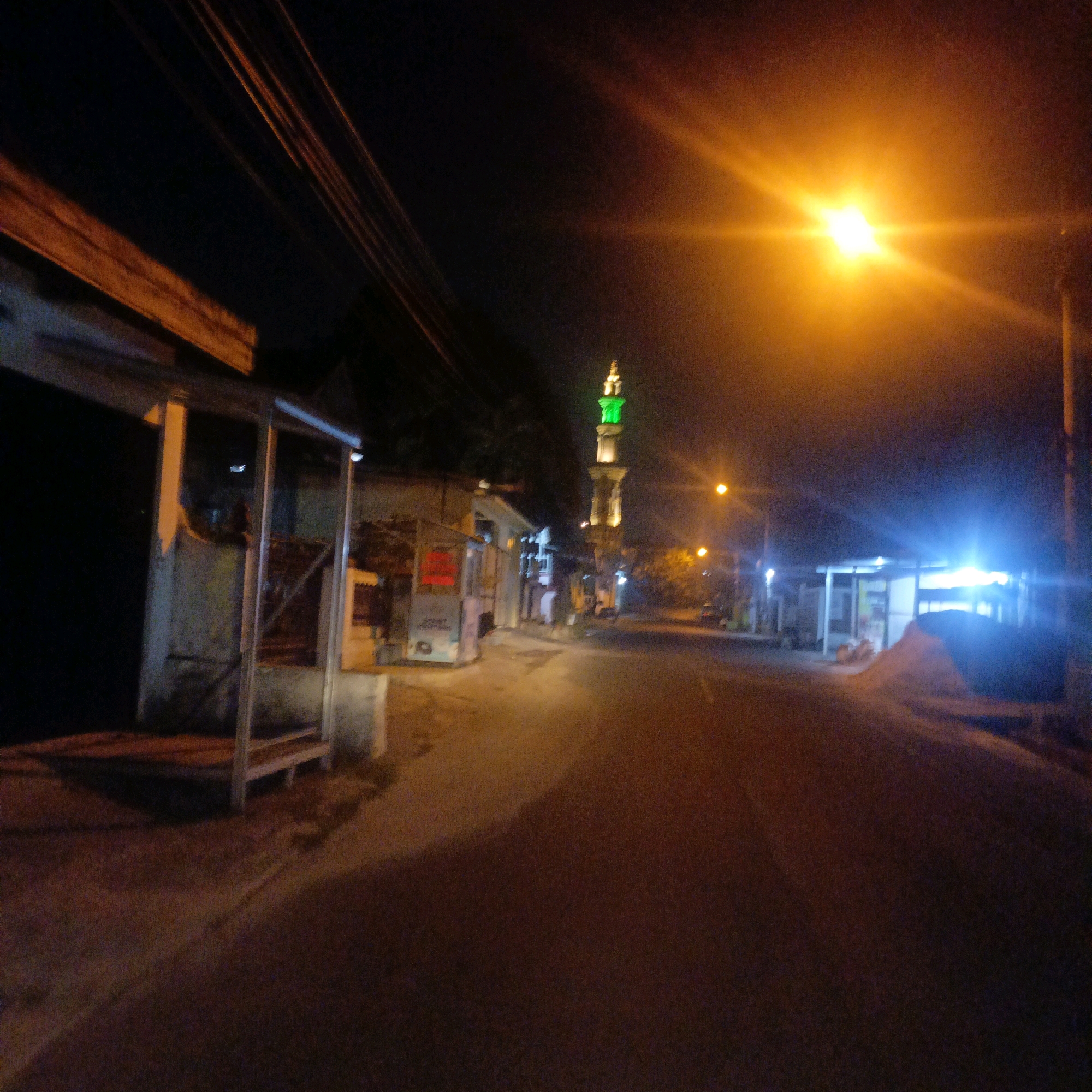 This image depicts a night scene on a road in Salatiga, Indonesia, characterized by low light conditions and artificial illumination. The focal point on the left is a simple, open-sided structure with a wooden roof and metal supports, possibly a bus stop or shelter. Further down the road, a mosque minaret with a green illuminated top stands prominently against the dark sky. Several streetlights cast a strong orange glow, with one particularly bright, overexposed light dominating the right side of the frame. The road surface appears to be asphalt, with some scattered debris or sand visible. Buildings line the left side of the road, with some appearing to be small shops or residences. The overall impression is one of a quiet, possibly suburban, street at night, with the mosque serving as a significant landmark.