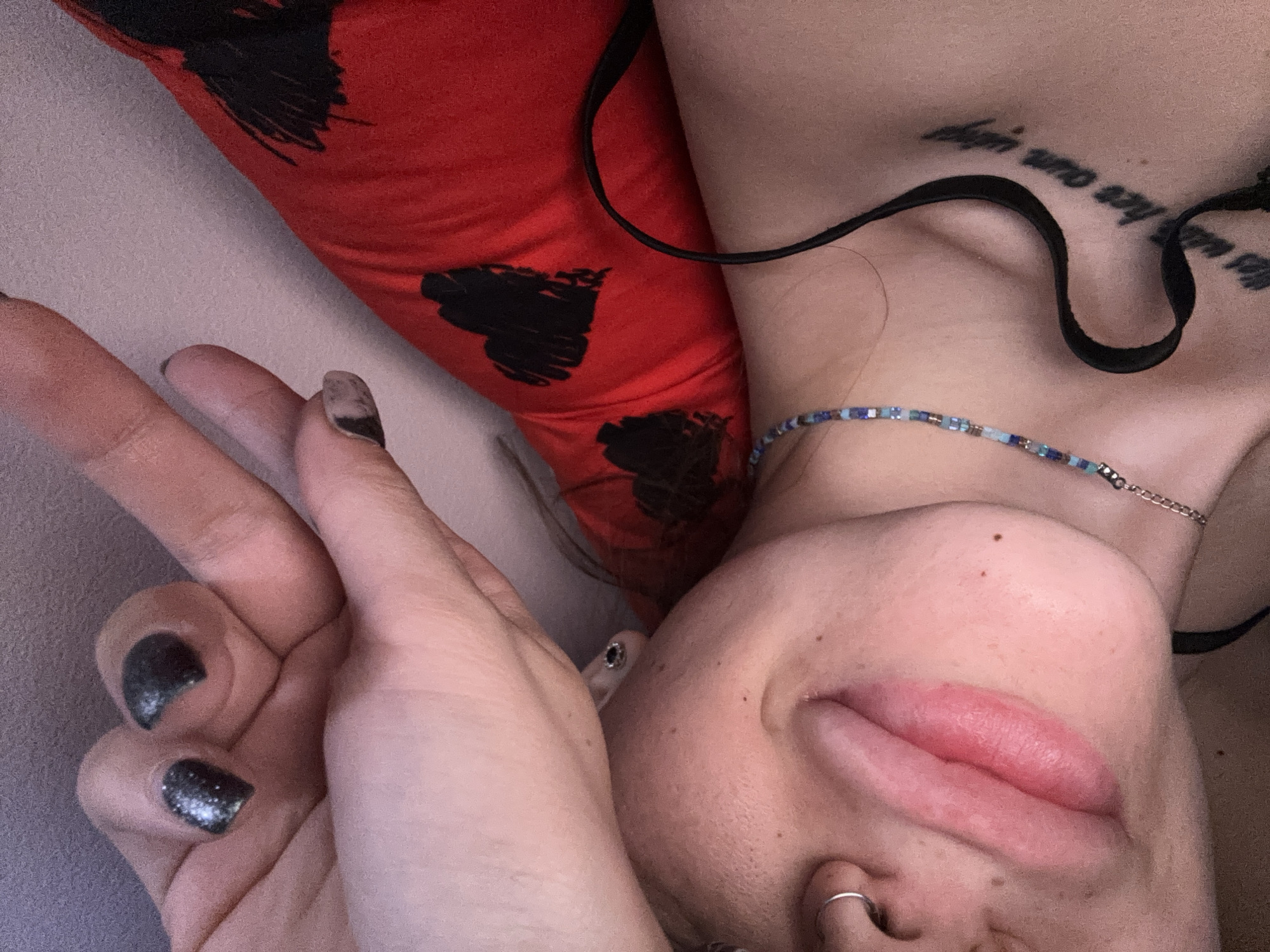 A person is lying on their back with a red pillow, showing a peace sign with their hand. They have dark nail polish and a nose ring. A tattoo is visible on their chest. The scene does not definitively show a location in Sofia, Bulgaria, nor does it allow to verify if the person is in Bulgaria.