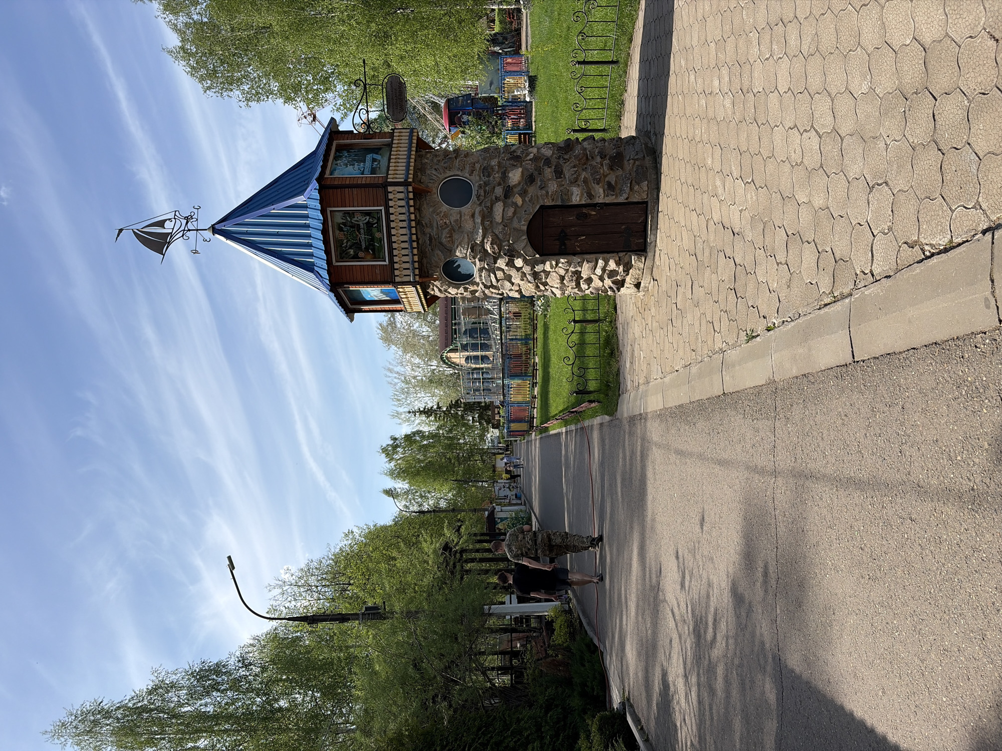 The image shows a stone tower with a blue roof and a weathervane on top, situated beside a paved pathway in a park-like setting. The tower has a wooden section with windows displaying images. A cobblestone path leads up to the tower's entrance, bordered by a sidewalk and a grassy area with a fence. Two individuals are walking on the pathway, further down the path there are other people and colourful playground equipment visible in the background, suggesting an amusement area within the park. The sky is light blue with scattered clouds, and trees line the path.