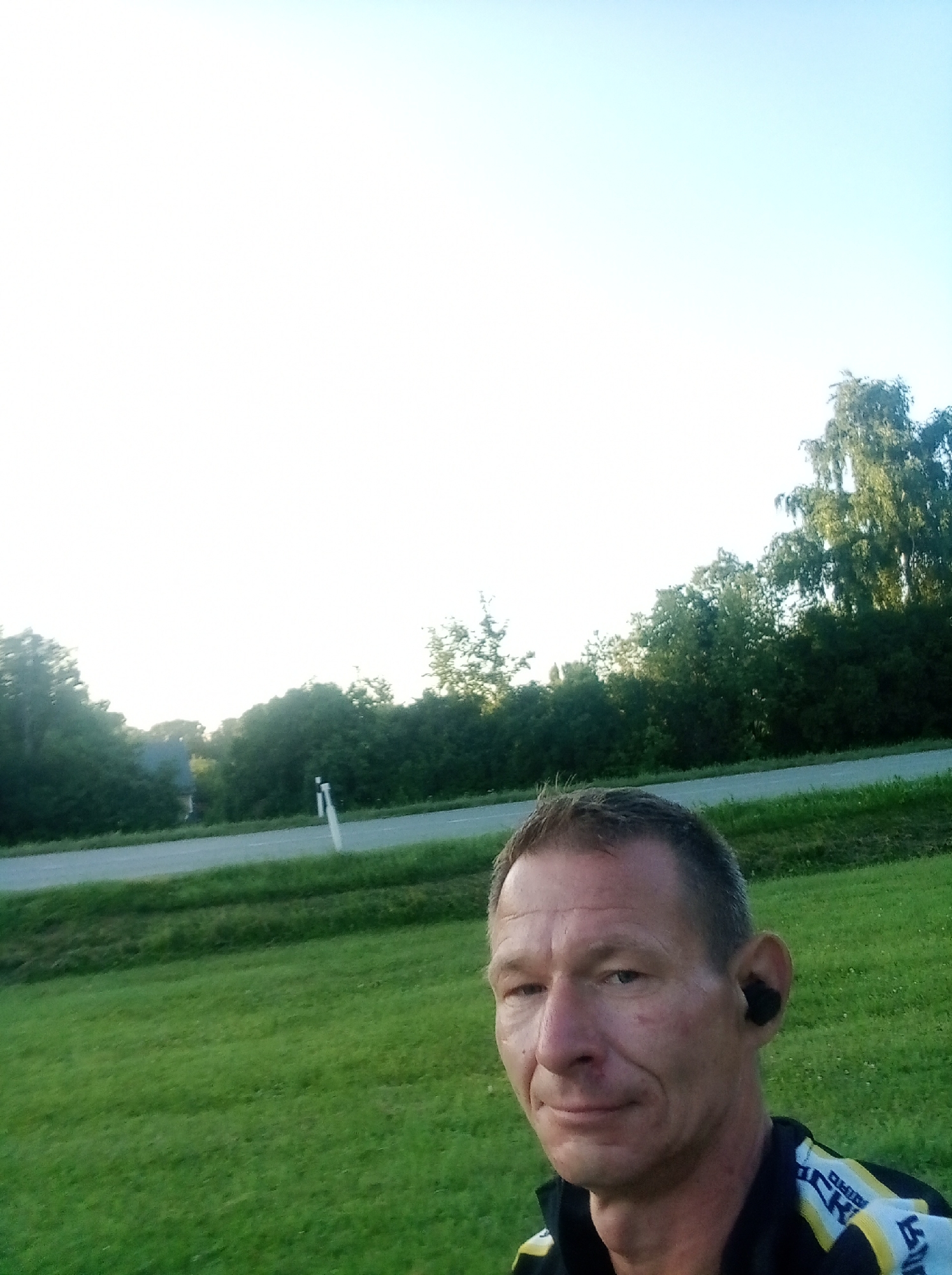 A medium-shot, selfie-style photograph shows a middle-aged man with short, graying hair and a light complexion. He is wearing a black jacket with yellow and white accents, and black wireless earbuds in his right ear. The man is outdoors in a grassy area, with trees and a road visible in the background. The sky is clear and bright. The overall scene suggests an outdoor activity such as running or cycling, possibly in a rural or suburban setting within the city of Tapa, Estonia, given the presence of trees and a paved road with a white marker post.