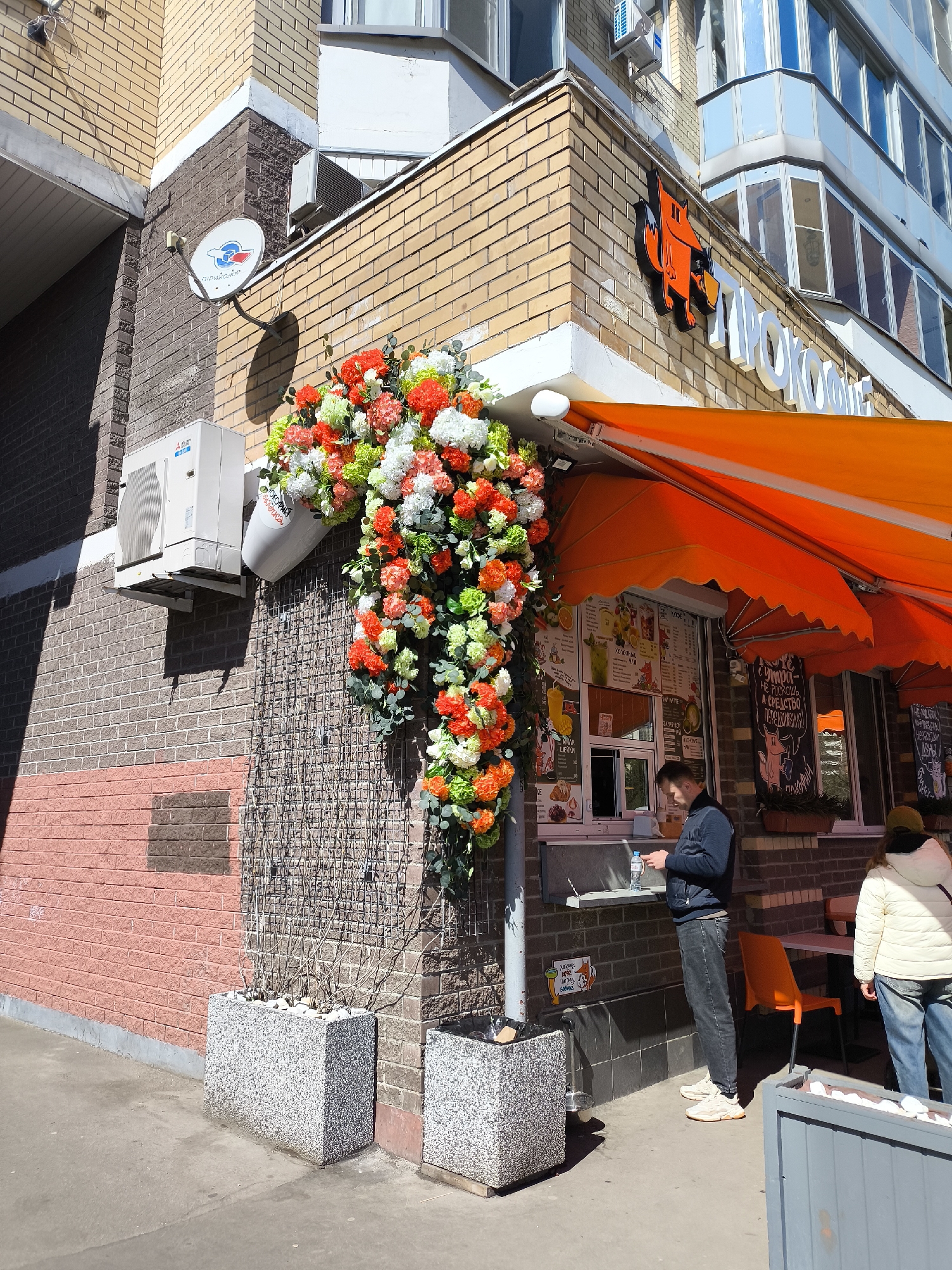 This is an outdoor shot of a corner building, likely a café or shop, in Moscow, Russia. The building's corner features a decorative display of orange, white, and green flowers cascading from a white container, affixed to a wire mesh background. Below this floral arrangement are two grey-speckled planter boxes. To the left, there is an air conditioning unit and a satellite dish mounted on the brick wall.
On the right side of the corner, there is an orange awning over a window where a man is standing, looking at his phone. He is wearing a blue jacket, grey jeans, and light-colored sneakers. Another person, wearing a white jacket and jeans, is visible to the right, partially obscured by the edge of a grey fence. An orange chair is positioned near them.
Above the awning, there is a sign with an orange fox design and Cyrillic text that translates to "PROKOFY", which is likely the name of the café. The building behind the café has a light blue hue with multiple windows. The environment is bright and sunny.
