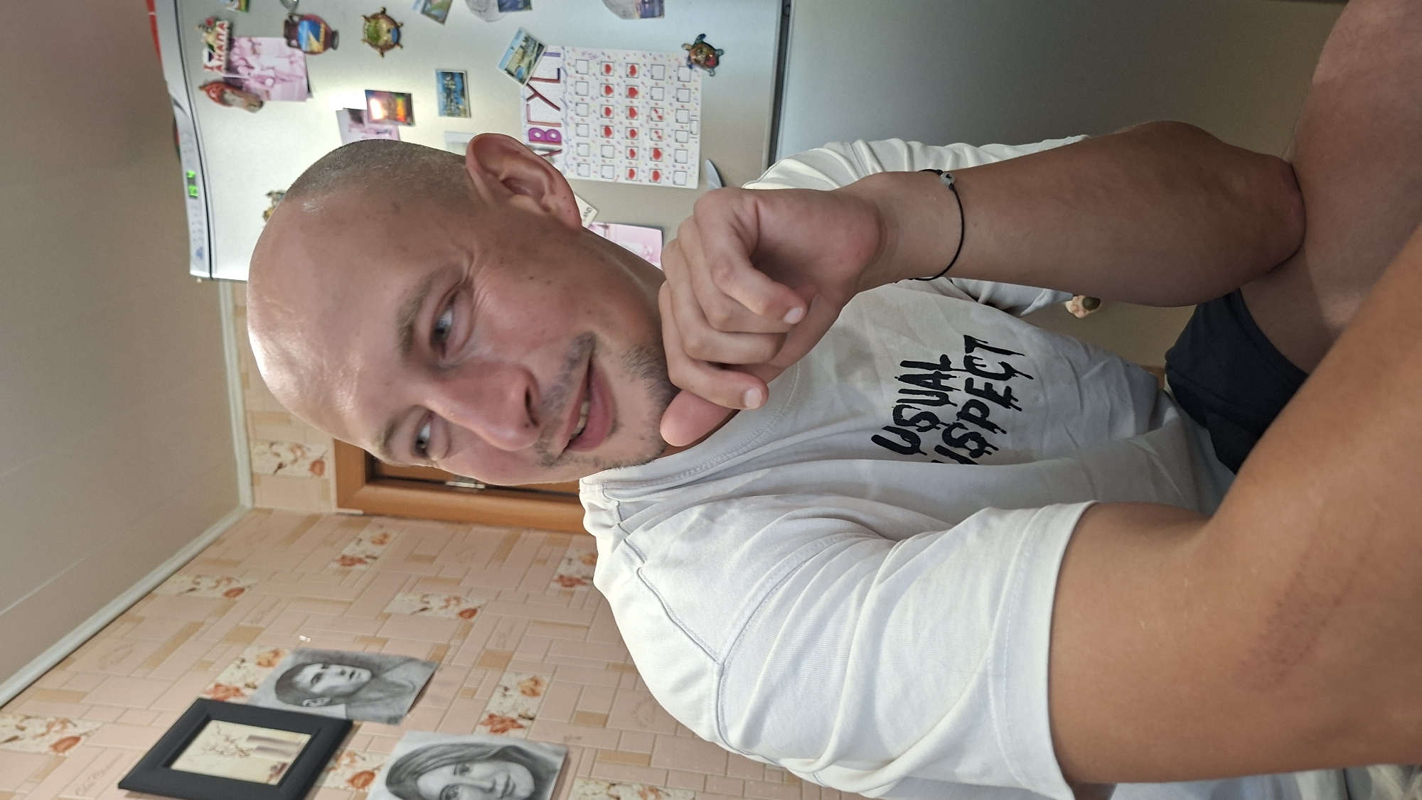 A medium shot captures a bald, light-skinned man with a short beard and light eyes, looking to his left with a slight smile. He is wearing a white long-sleeved t-shirt with black lettering that reads "USUAL INSPECT" and dark blue shorts. The man is seated, with his right arm resting on his knee and his chin propped on his left hand. The background features a light pink tiled wall adorned with floral patterns. To the left, a framed black and white portrait of a man hangs on the wall. To the right, a silver refrigerator is visible, decorated with various magnets and a calendar. The overall setting appears to be an indoor domestic environment, possibly a kitchen, in Myadzyel, Belarus.