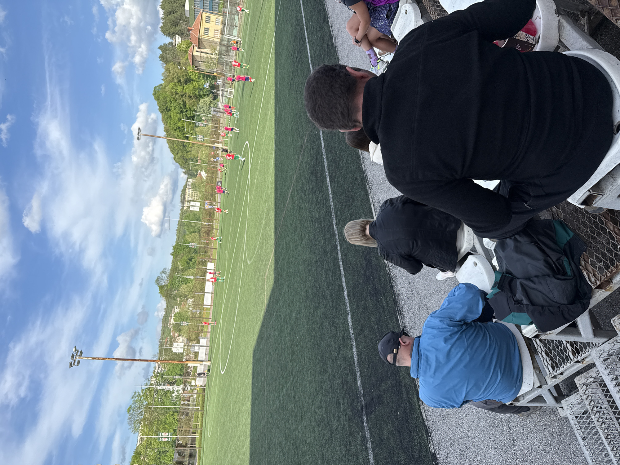 The image shows an elevated view of a soccer field. People sit on stadium bleachers and watch a soccer game. Players in green and red uniforms are visible on the field. There are light posts, and buildings. The scene is set in Gabrovo, Bulgaria.