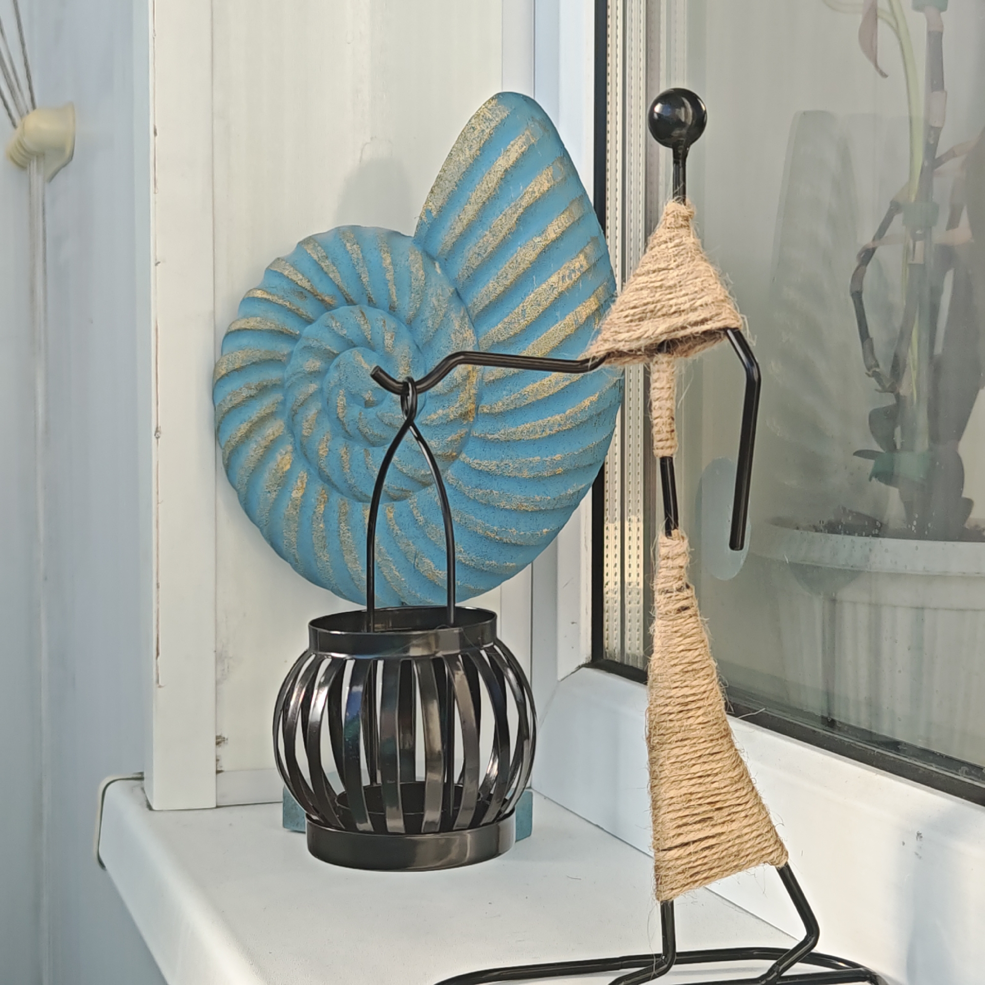 On a white windowsill in Rostov, Russia, a still life is arranged. From left to right: a white electrical outlet, a blue-and-gold spiral seashell artifact, a black wrought-iron hanging lantern, and a black metal wire figure wrapped in jute twine. Through a nearby window, a potted plant is visible.