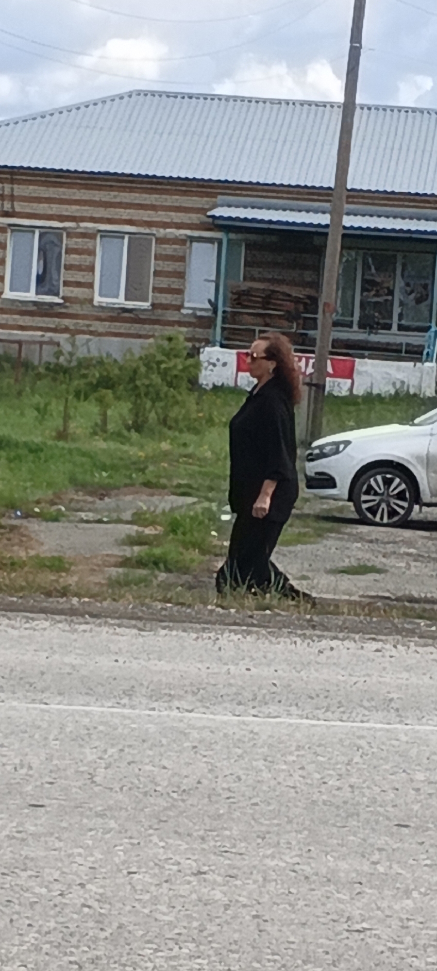A woman with dark, wavy hair and sunglasses is walking on a grassy area next to a paved road. She is wearing a black top and black pants. In the background, there is a building with a corrugated metal roof and striped brick walls, as well as a white car. A utility pole stands to the right of the woman. The overall scene appears to be in a rural or suburban area, consistent with the city of Kargat, Russia.