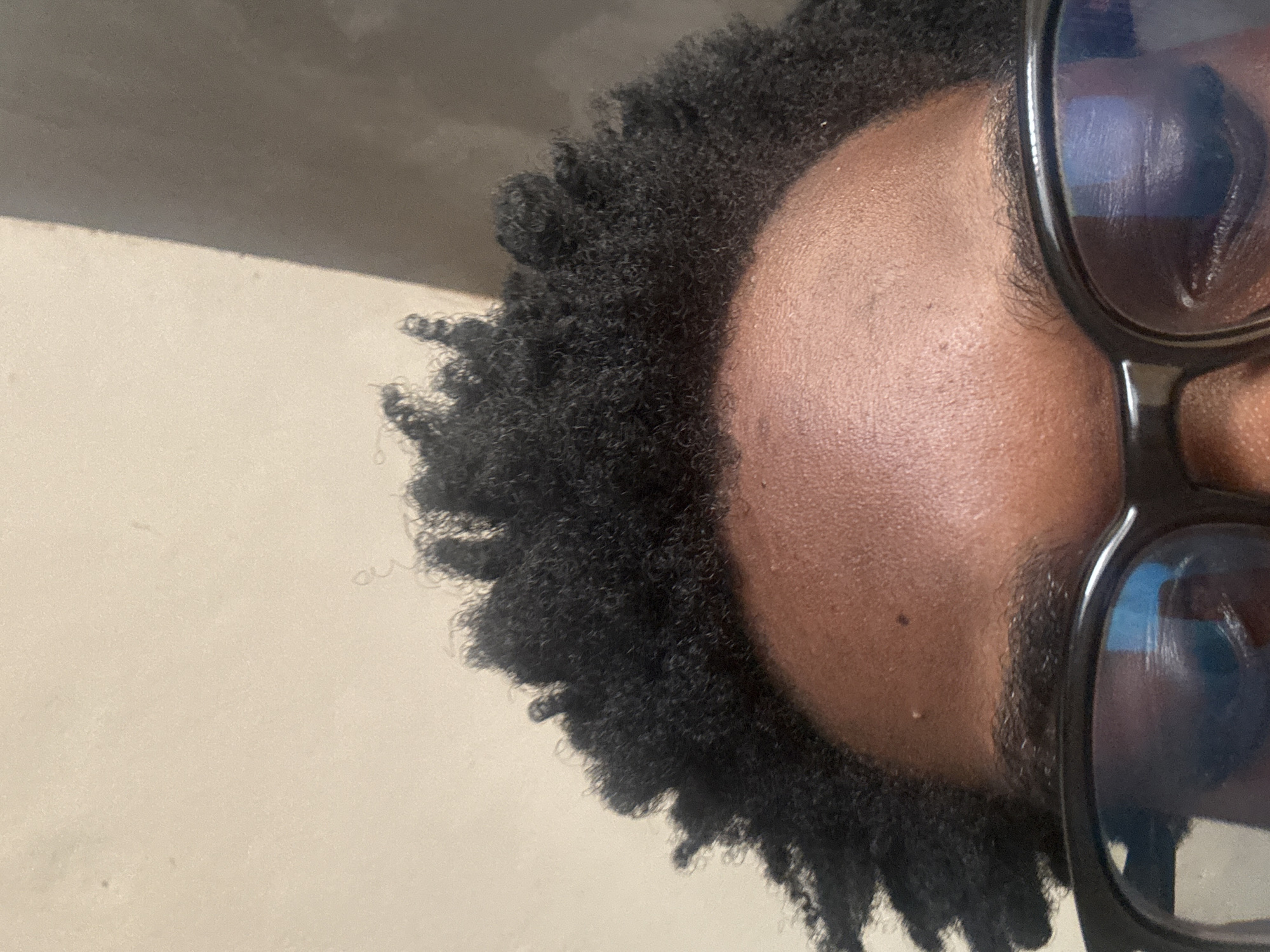 Close-up image of a person's head and face, wearing glasses with reflective lenses. The person has dark, tightly curled hair. The skin tone is brown. The image appears to be rotated. The background is a plain, light-colored wall. No specific location in Ihiagwa, Nigeria can be determined from the image.